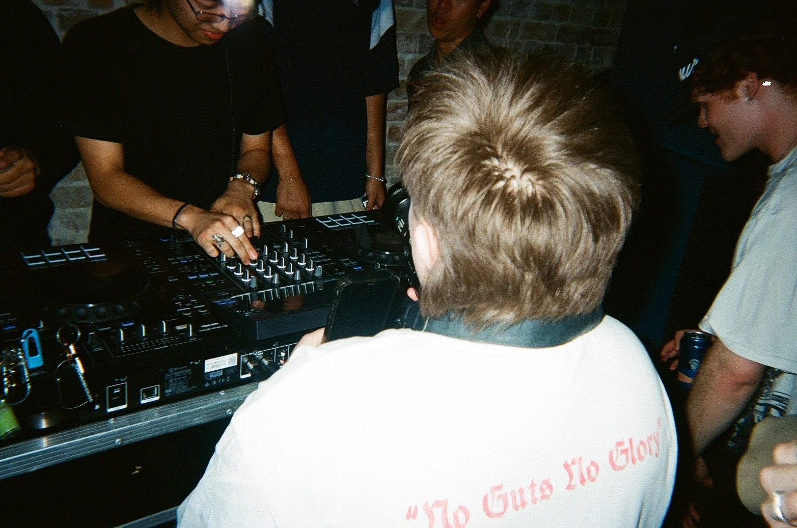 People gathered around a DJ mixing music with a DJ controller at a party or club, with some individuals holding drinks.