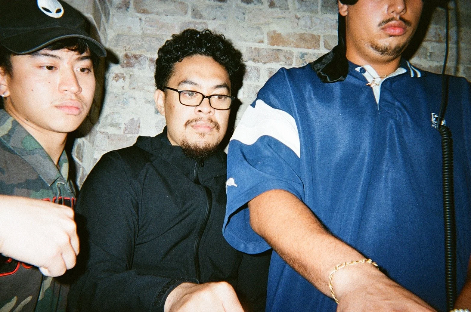 Three men standing close together indoors against a brick wall, with the focus on the middle man wearing glasses and a black jacket.