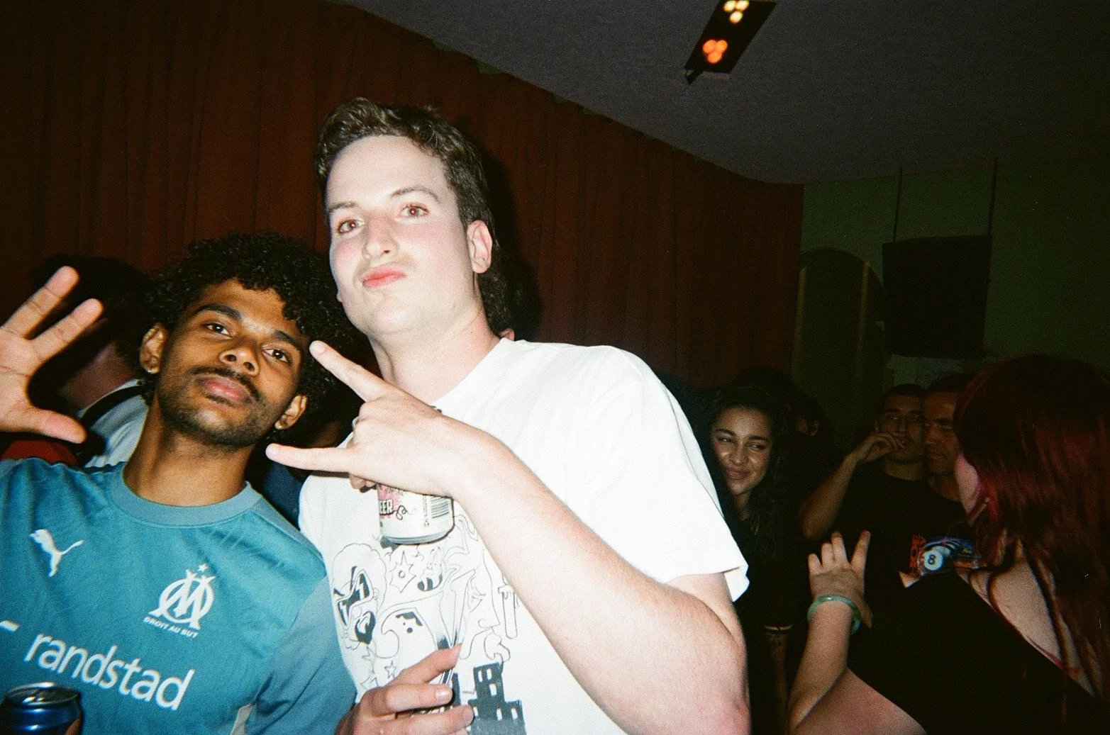 Group of young people partying at a nightclub, two men in the foreground making peace signs, and others in the background enjoying the event.
