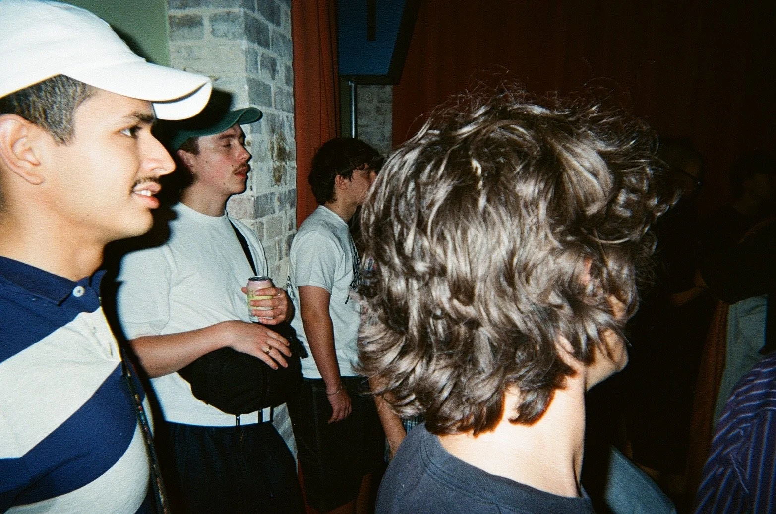 Line of young men standing indoors against a brick wall, some wearing hats and casual clothing, with one holding a drink, in a possibly social setting.