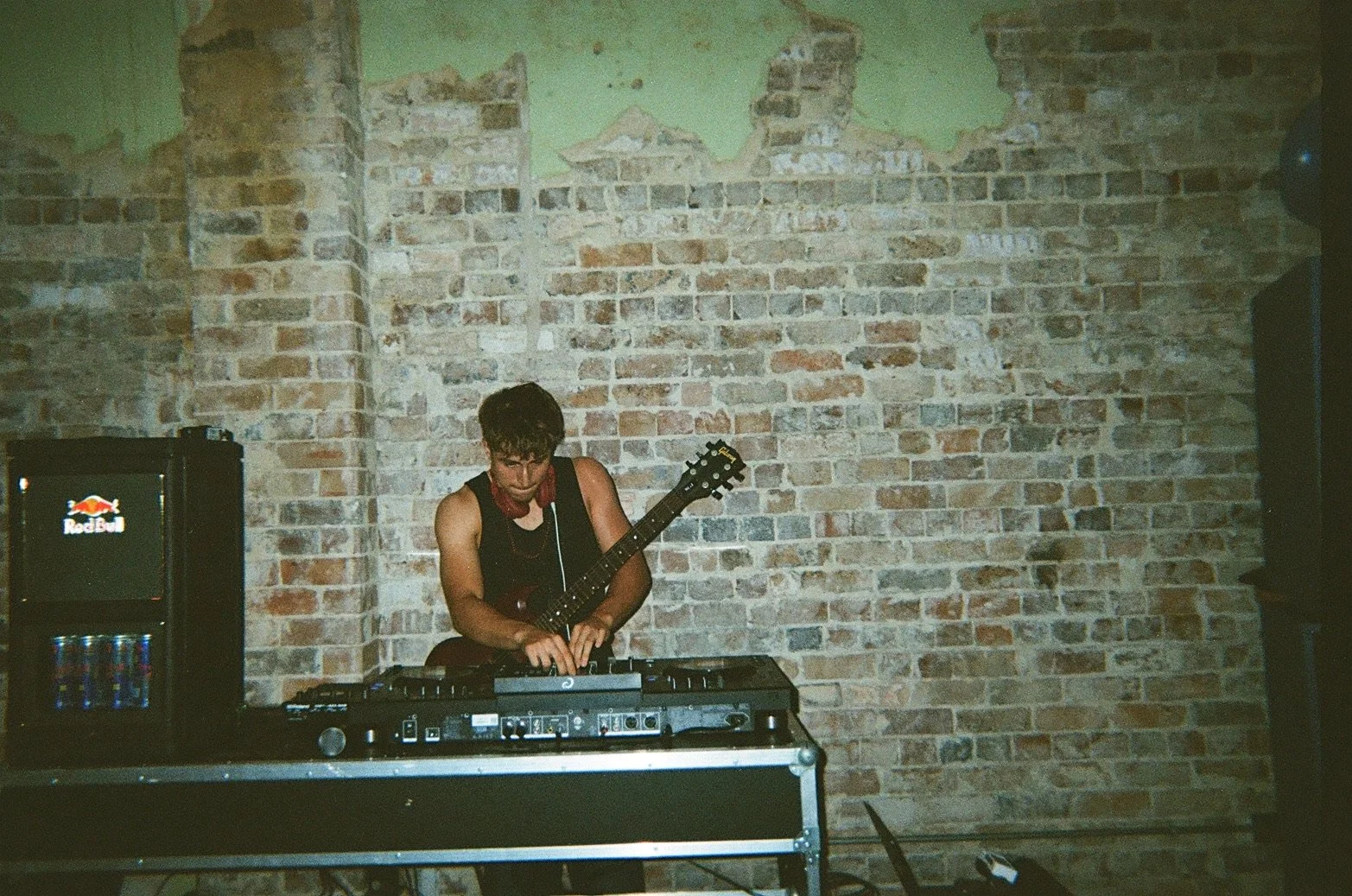A male DJ is performing in front of a brick wall, using a mixer and a keyboard, with a Red Bull refrigerator to his left.
