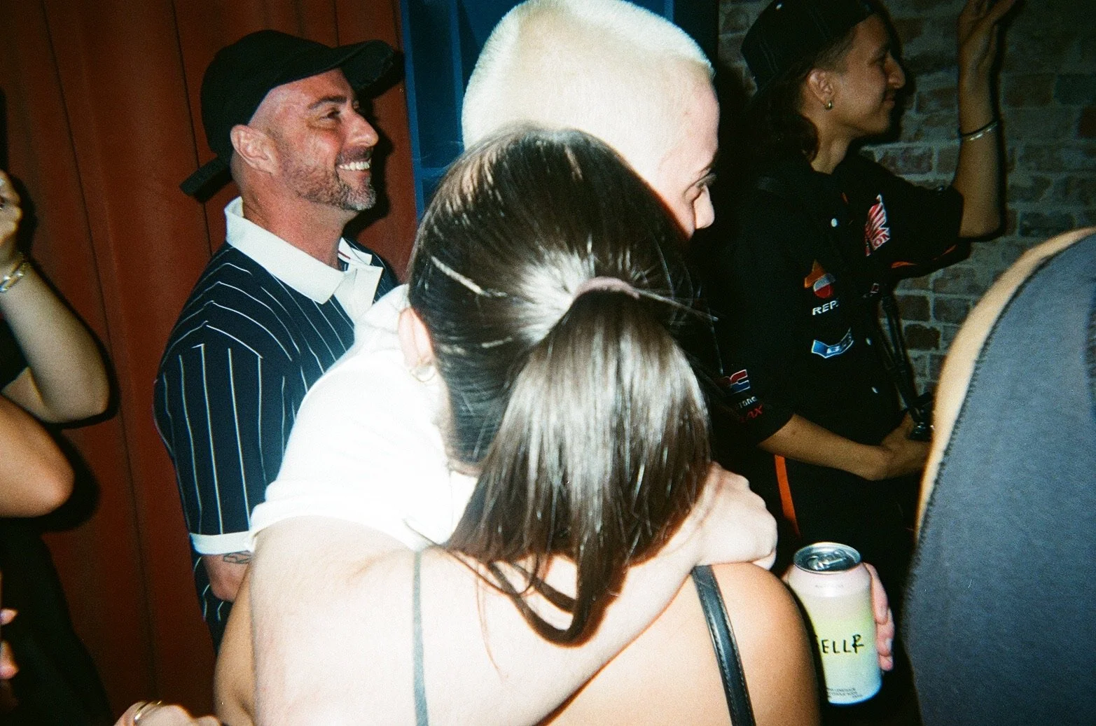 Group of people at a social gathering, some smiling, one holding a can of drink.