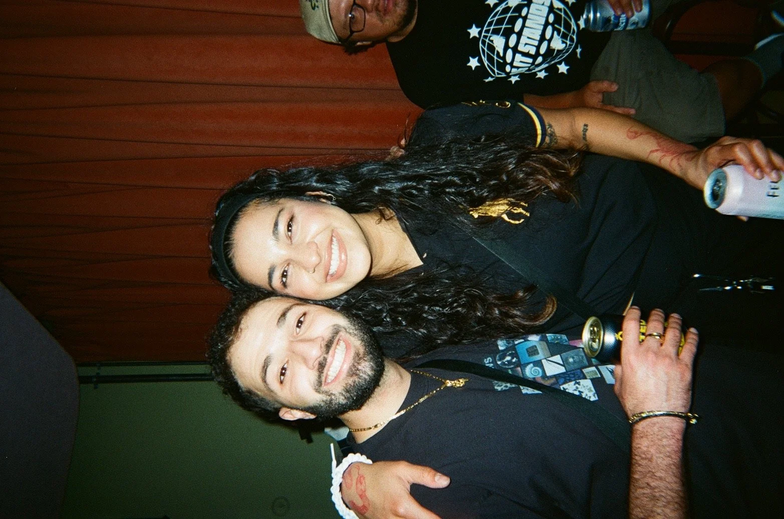 A man and a woman smiling, sitting together, each holding a drink, in front of a wooden wall. The woman has long dark wavy hair and is wearing a black shirt, and the man has short dark hair, a beard, and is wearing a black t-shirt with a colorful des