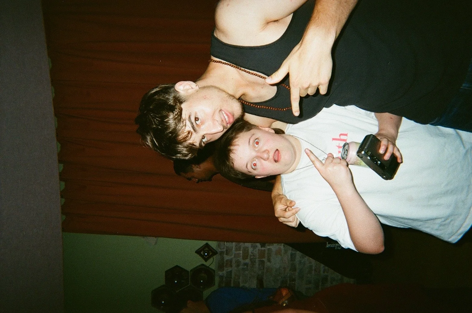 Two young men posing playfully together; one is pointing at his friend’s chest, and the other is making a face while holding a device in his hand. They are in front of a green wall with a brick pattern at the bottom and a background of closed brown c