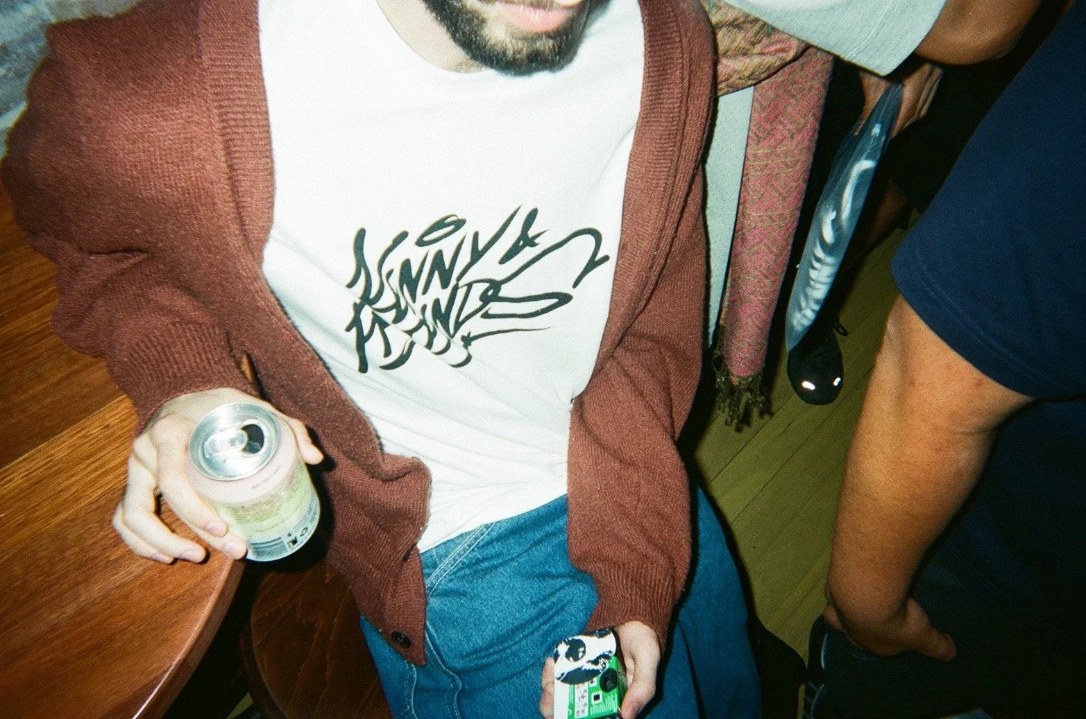 A person is sitting at a wooden table holding a can of beverage and a phone. They are wearing a white T-shirt with black scribbled text, a reddish-brown cardigan, and blue jeans. Another person is standing nearby, wearing dark shorts, and a small obj