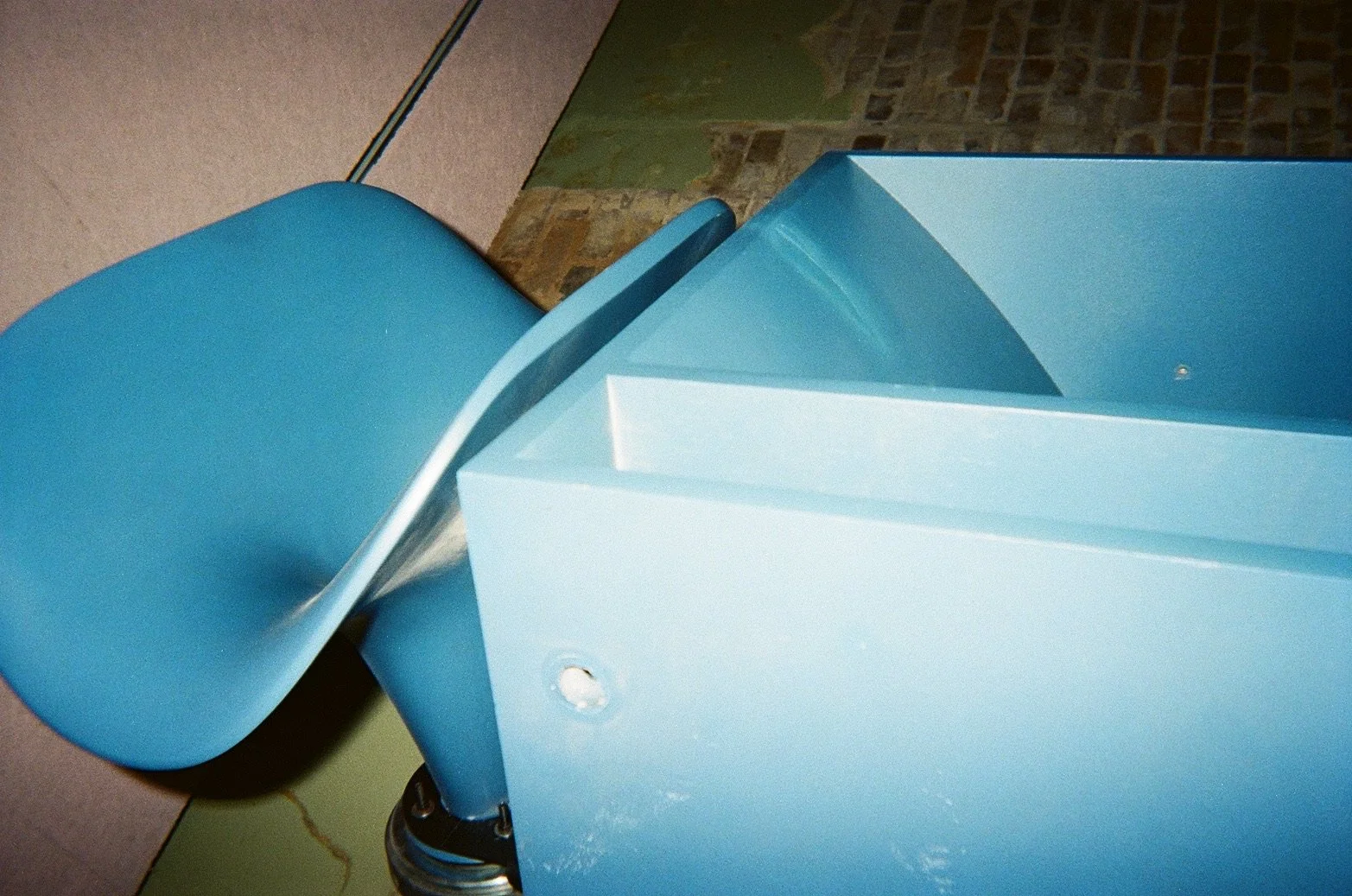 A blue vintage barber's chair with a rounded back and armrests, partially visible in an indoor setting with a tiled wall and a greenish part of the wall nearby.