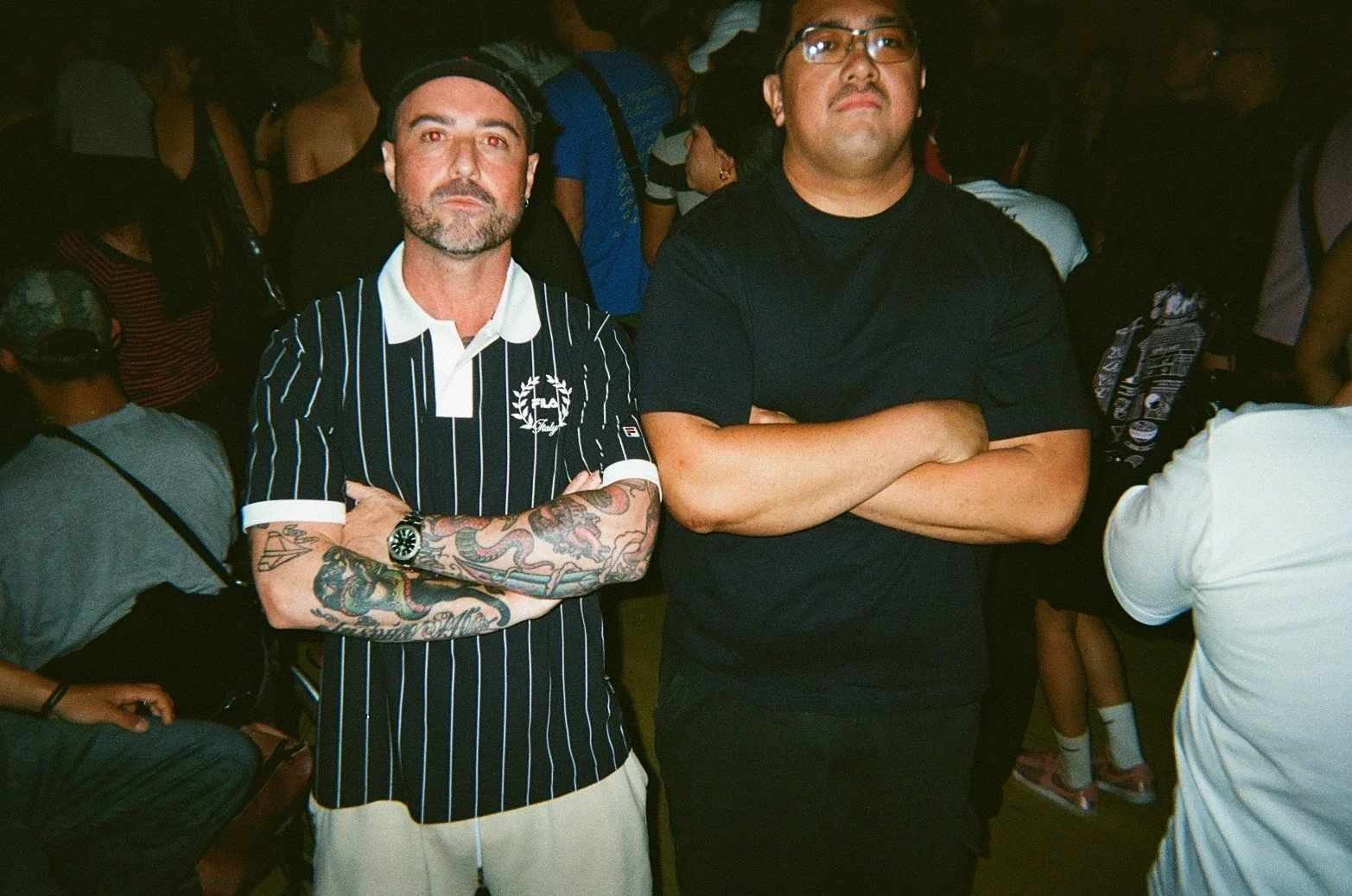 Two men standing with arms crossed at a crowded indoor event. The man on the left has tattoos on his left arm, a beard, and is wearing a black and white striped polo shirt. The man on the right has glasses, a serious expression, and is dressed in a b