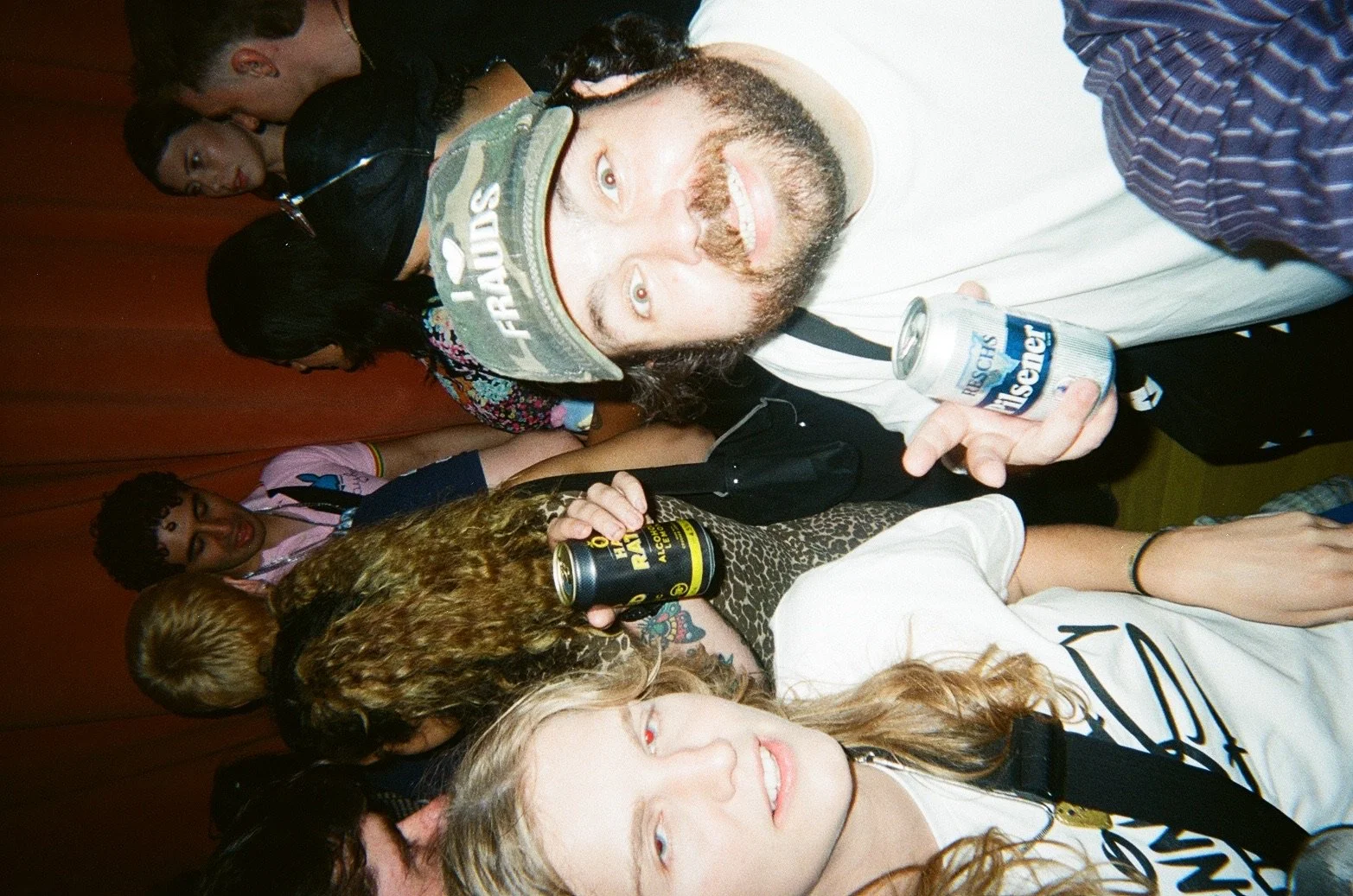 Group of people at a social gathering, some holding cans of beer, in an indoor setting with a wooden wall background.