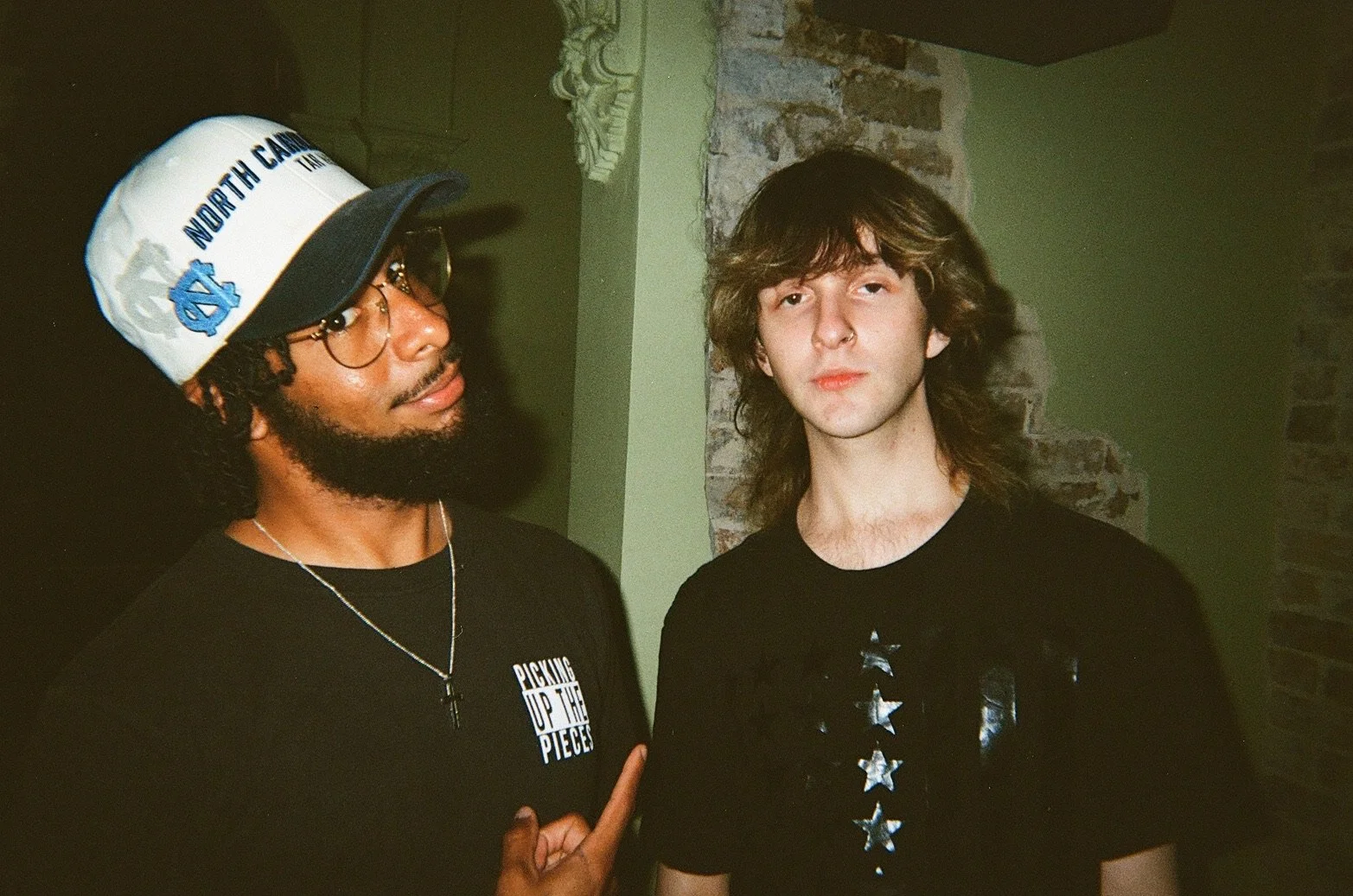 Two men standing indoors in front of a brick and light green wall. The man on the left has dark skin, curly hair, glasses, a white North Carolina cap, and a black t-shirt that reads 'Picking Up The Pieces'. The man on the right has light skin, long w