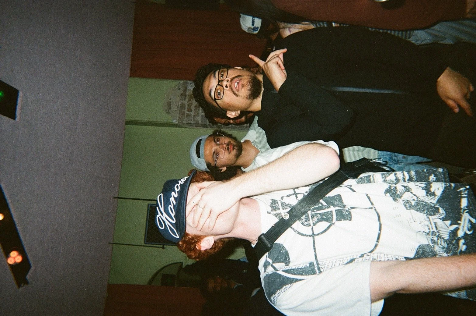 A man with red hair and a beard wearing a  baseball cap and colorful graphic t-shirt is wrapping his arm around a woman with curly hair, who is touching his face, in a lively indoor setting with green walls and a wooden partition.