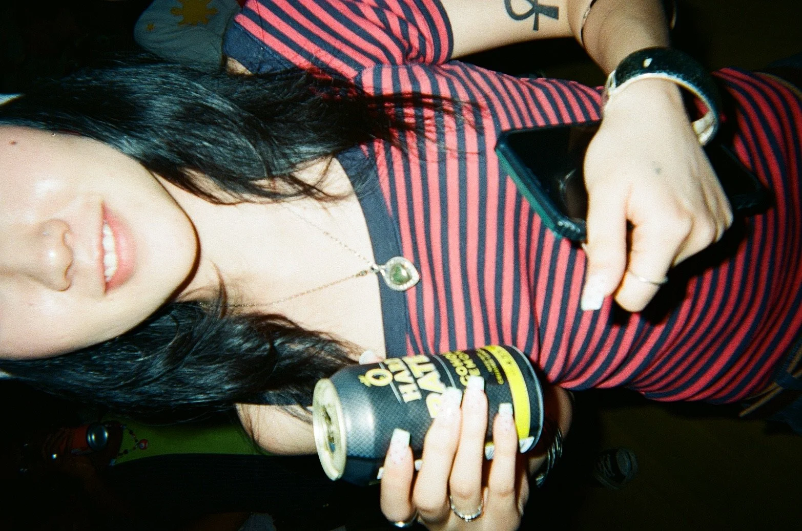 A woman with black hair and light skin, wearing a red and navy striped shirt, holding a can of energy drink in one hand and a smartphone in the other, with a necklace and bracelet visible.