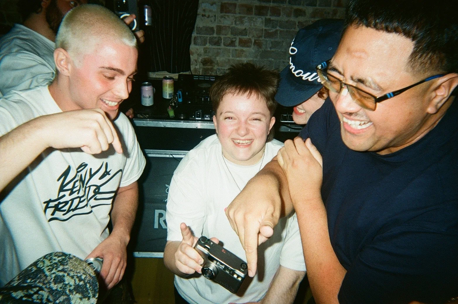 Group of young people smiling and laughing at a social gathering, with a DJ in the background and a person holding a camera in focus.