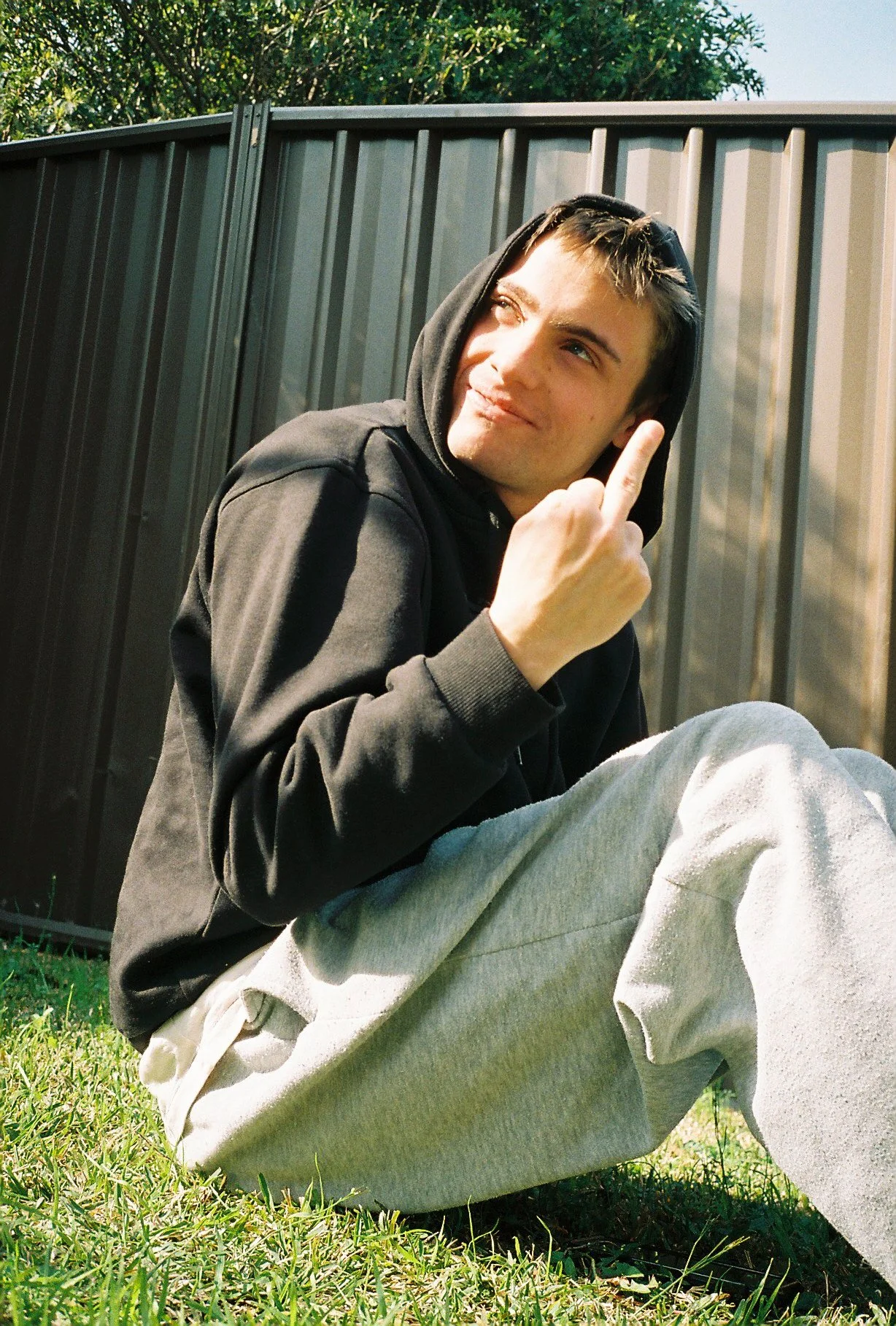 A young man sitting on grass in front of a metal fence, wearing a black hoodie and gray sweatpants, with sunlight on his face and hair.