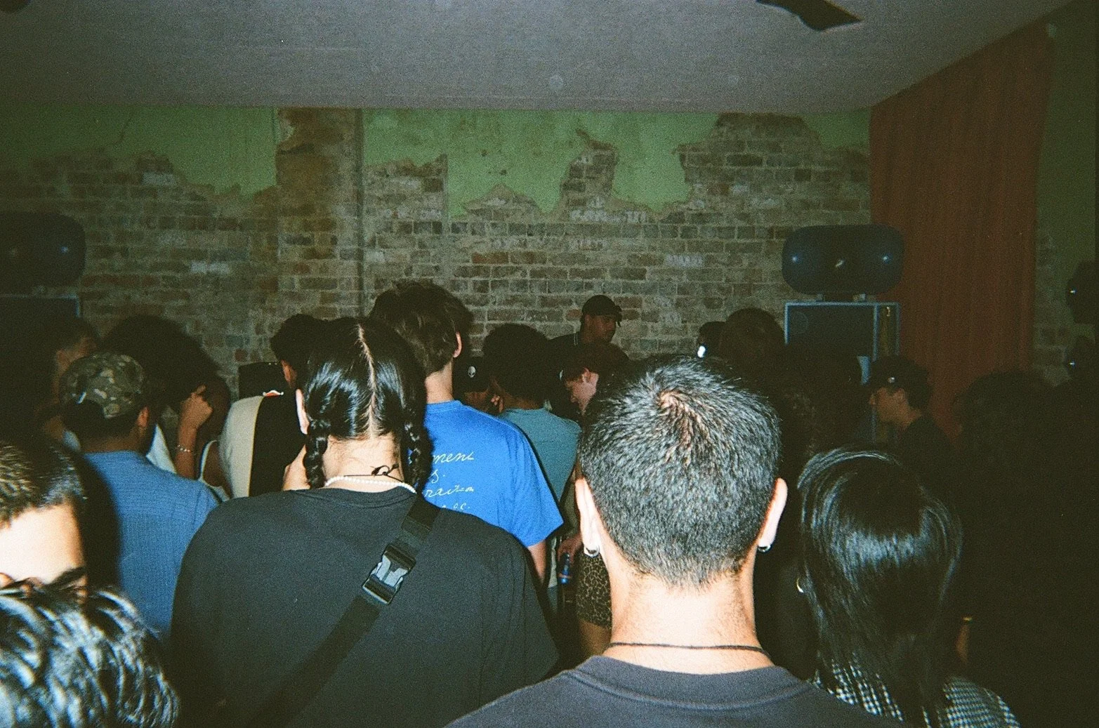 A crowd of people standing close together in a dimly lit room with a brick wall background, possibly attending a concert or event.