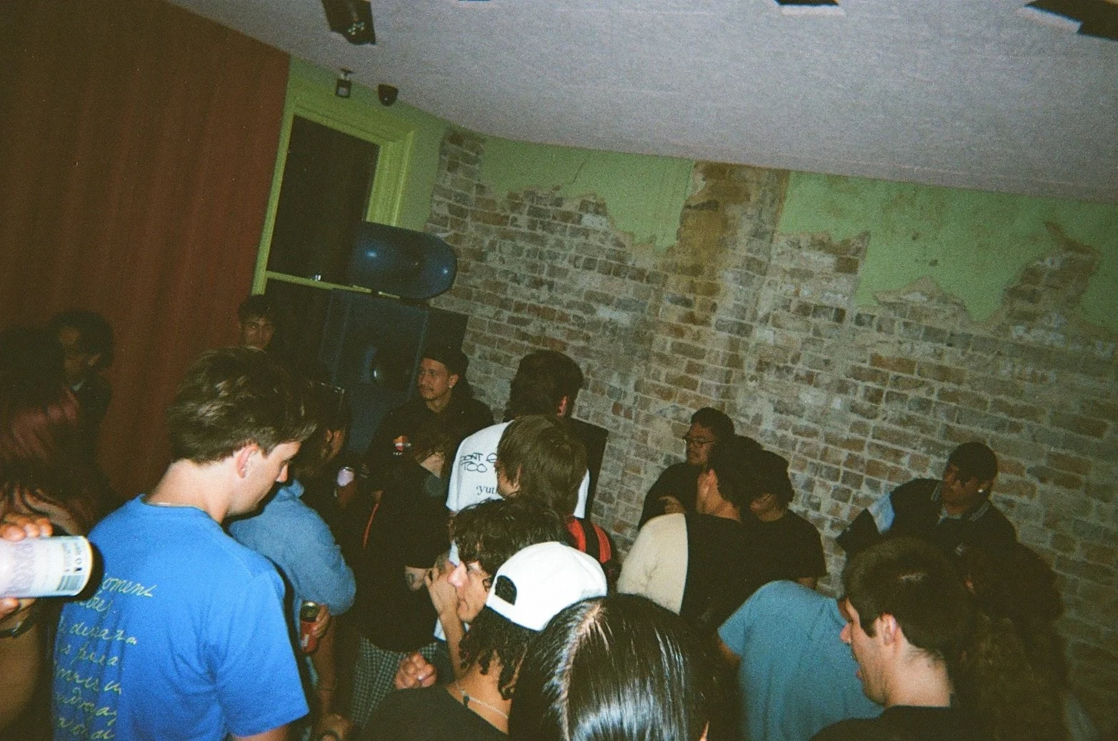 Young people gather in a dimly lit room with exposed brick walls and green paint, some are talking while others look toward a speaker at the front of the room, with a large speaker system nearby.