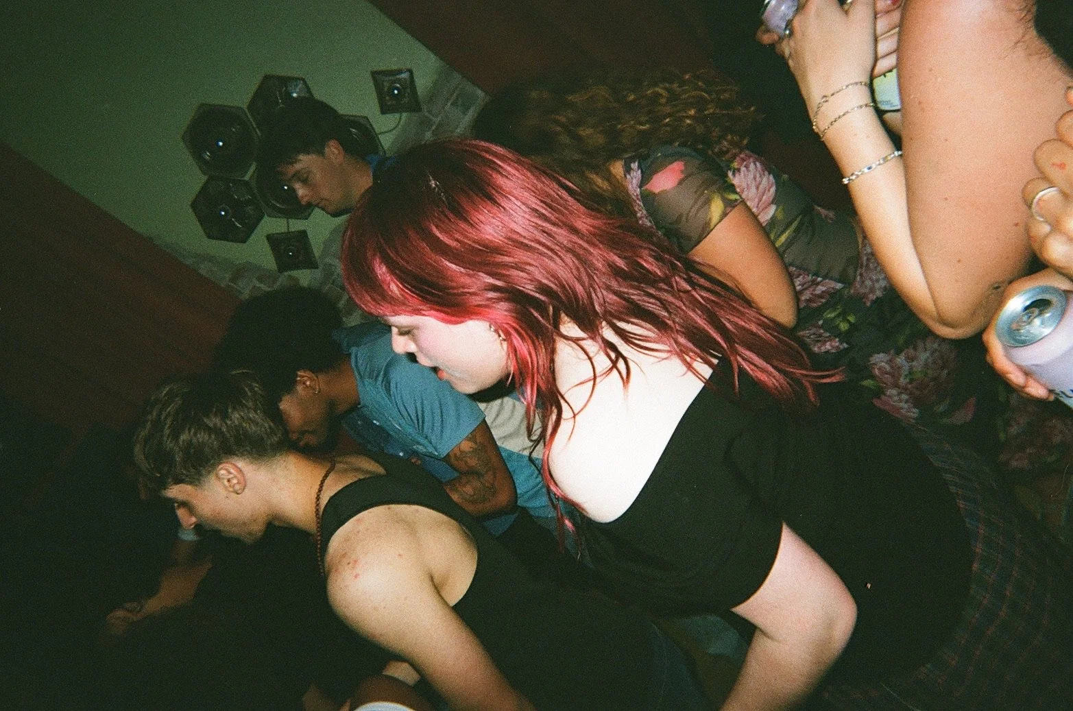 Group of young people dancing closely together in a dimly lit room, some holding drinks, with speakers mounted on the wall in the background.