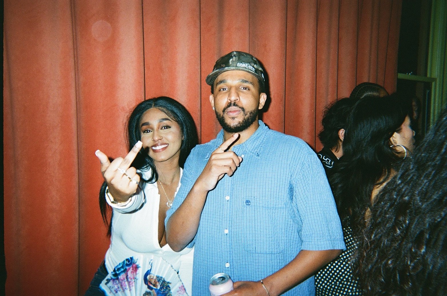 A group of people at a party, with a woman in a white top smiling and showing her middle finger, and a man in a blue shirt holding a drink, standing in front of a red wall.