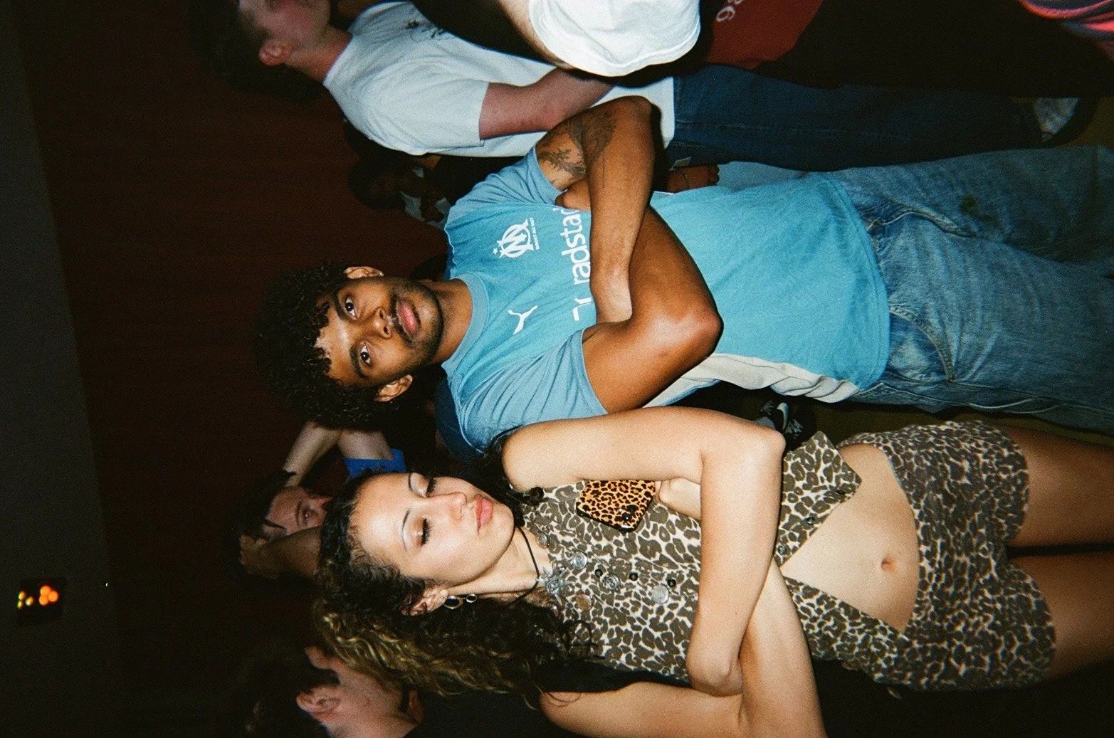 A young man with curly hair wearing a blue T-shirt with sponsor logos and denim shorts, standing with his arms crossed, looking at the camera. Beside him, a young woman with long curly hair, dressed in leopard print attire, with her arms crossed and 