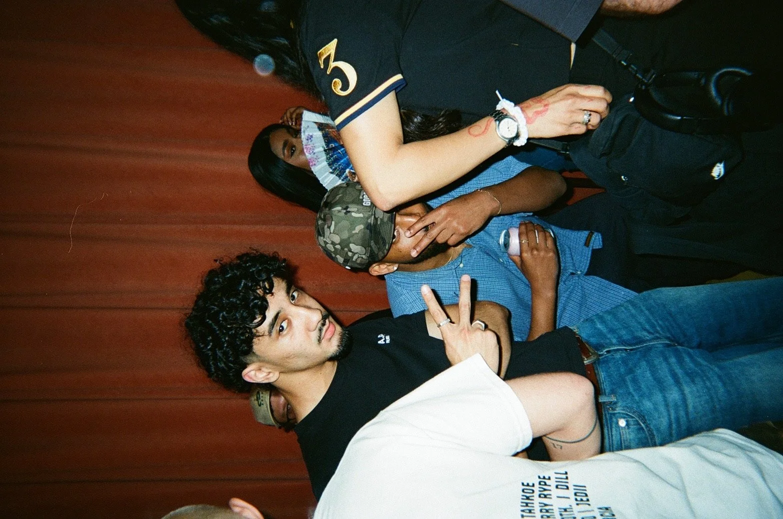 A group of five young people posing together in front of a red curtain. The man in front is making a peace sign with his right hand, has short curly hair, a black t-shirt, and jeans. Others are standing behind him, some wearing camouflage hats, masks