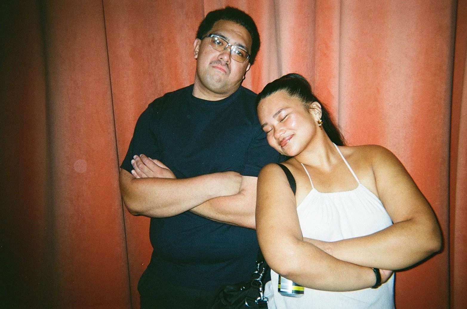 A man and woman standing side by side with their arms crossed, leaning against a red curtain. The man is wearing glasses and a black shirt, has short dark hair, and a serious expression. The woman has her eyes closed, is smiling, and wearing a white 