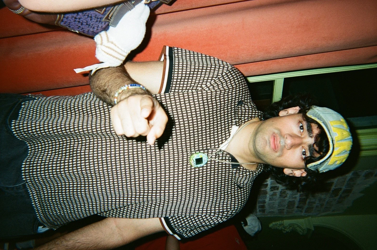 A young man with dark curly hair, wearing a Fish hat, a patterned shirt, a beaded bracelet, and a necklace with a green pendant. He is standing in a room with brick wall details, and pointing toward the camera.
