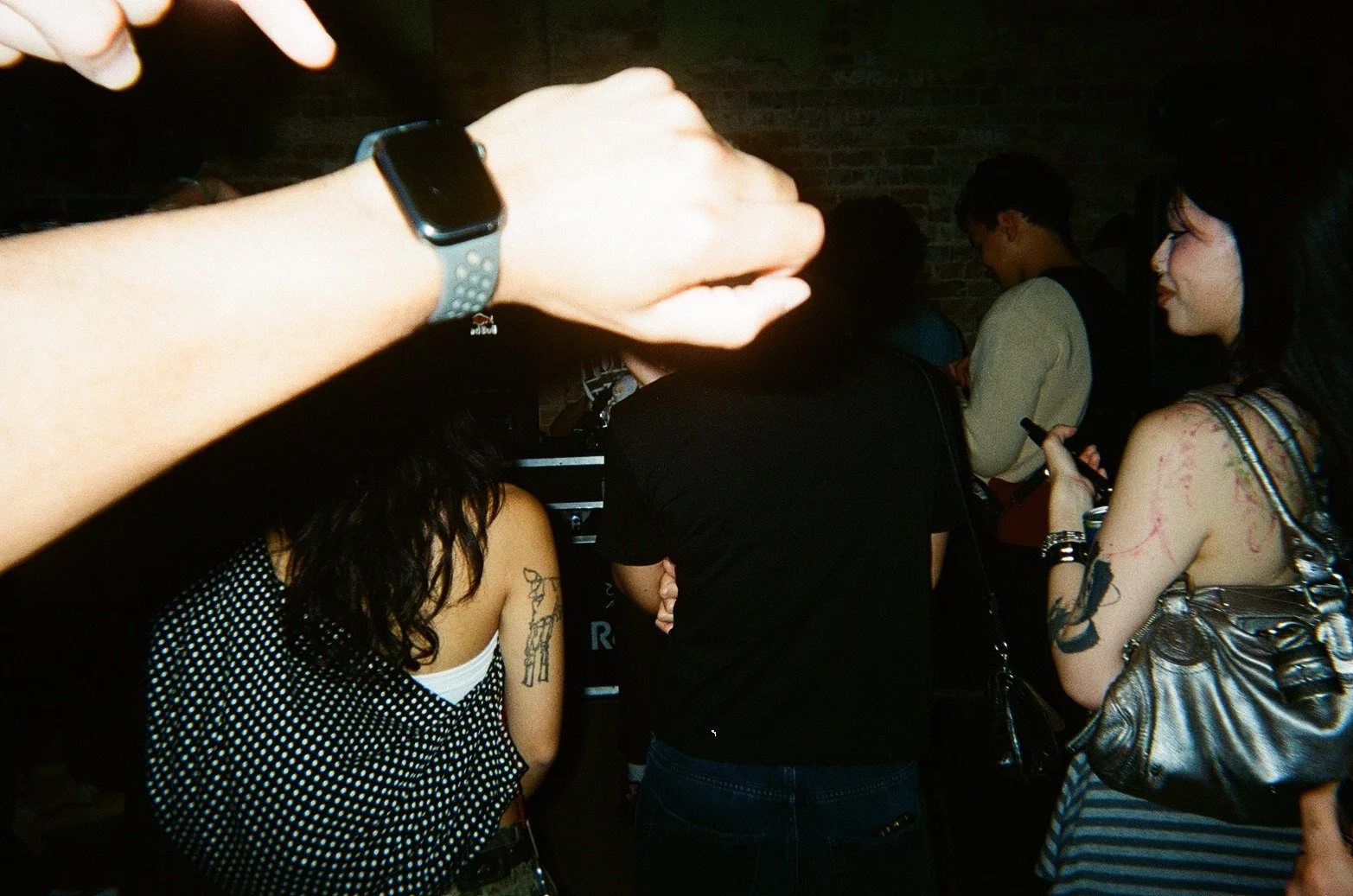 People gathered in a dimly lit indoor setting, some with tattoos, using their phones, and wearing casual clothing.
