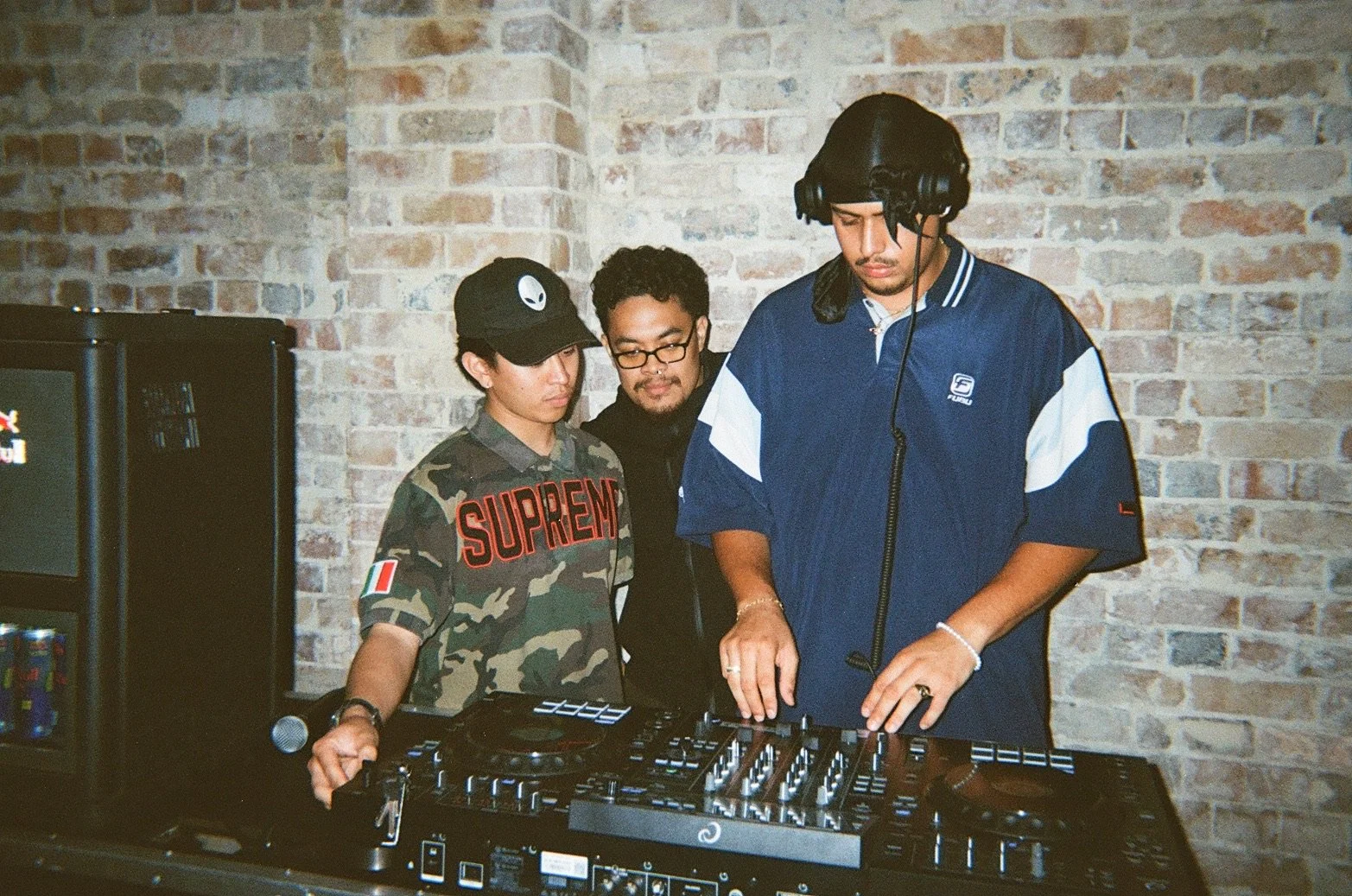 Three young men, one DJ wearing headphones, standing at a DJ booth with a brick wall in the background.