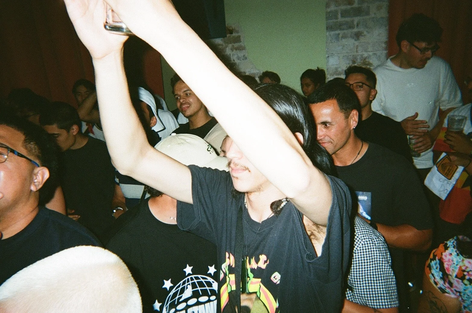 A group of people at an indoor event, with a man in the foreground raising his arm and holding a phone, surrounded by others engaged in conversation or observing, with brick and painted walls in the background.