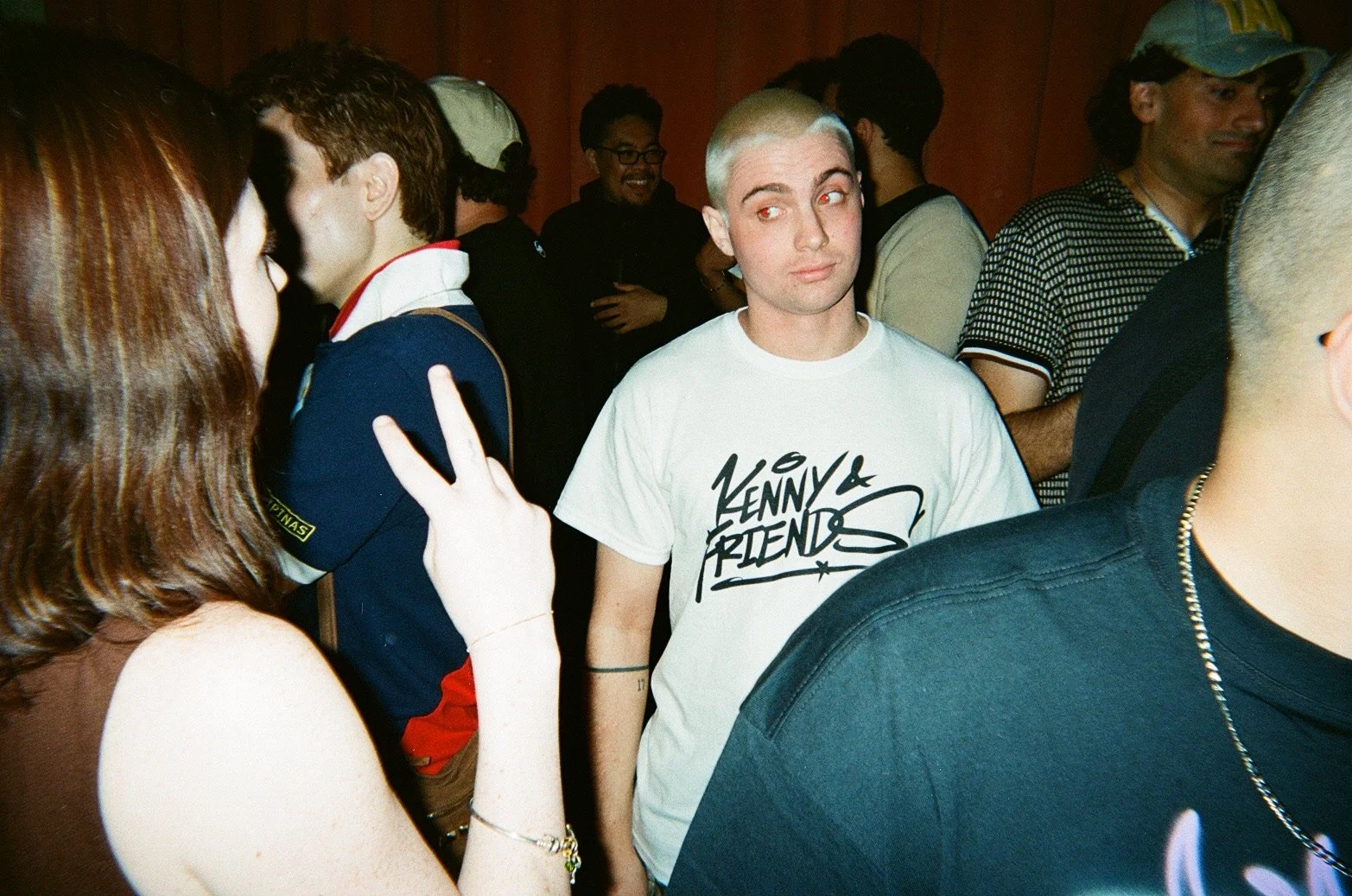 A group of people at a crowded indoor event, some engaged in conversation, and a man with bleached blonde hair and a white "Kenny & Friends" t-shirt standing in the center.