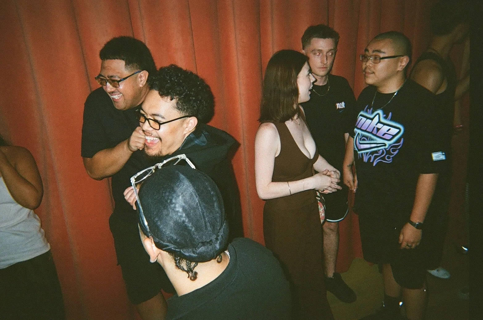 Group of five diverse young people socializing indoors, standing close together against a red wood-paneled wall, engaging in conversation and smiling.