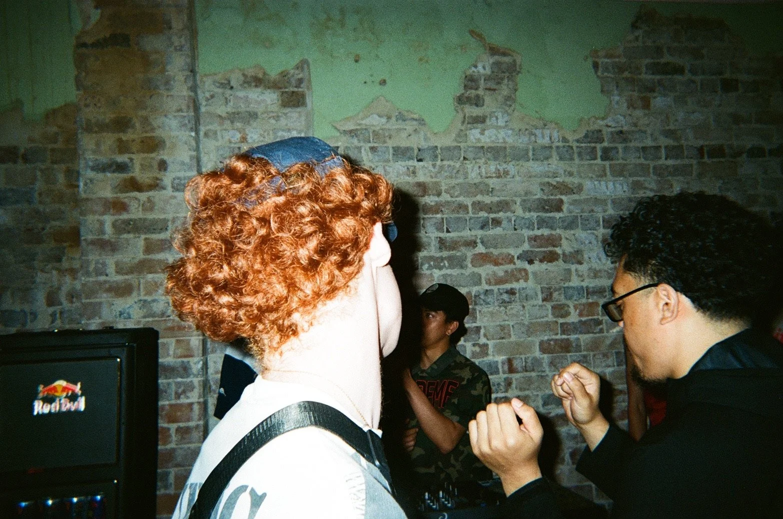 Person with curly red hair and a blue baseball cap facing another person with glasses. They are in an indoor setting with an exposed brick wall in the background.