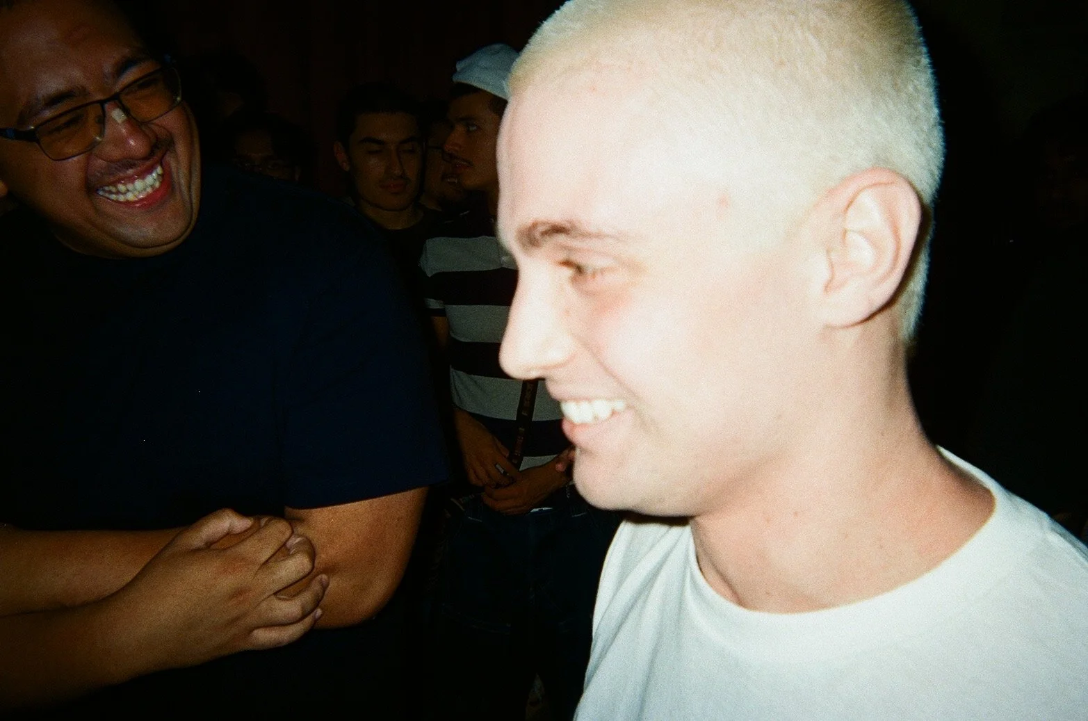 A group of people at a social event, with a young man with bleached blonde hair smiling and looking sideways in the foreground, and a man with glasses and a black shirt smiling in the background.