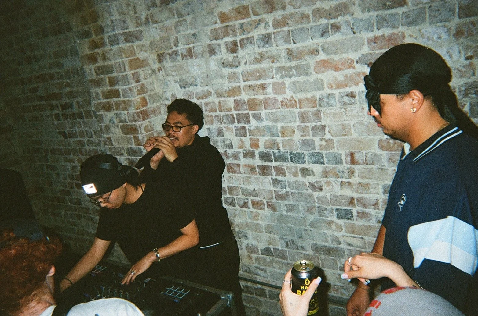 Three people at a party or social gathering, two men and one woman, against a brick wall. One man is holding a microphone and the woman is using a DJ controller, while the other man is holding a can of beer.