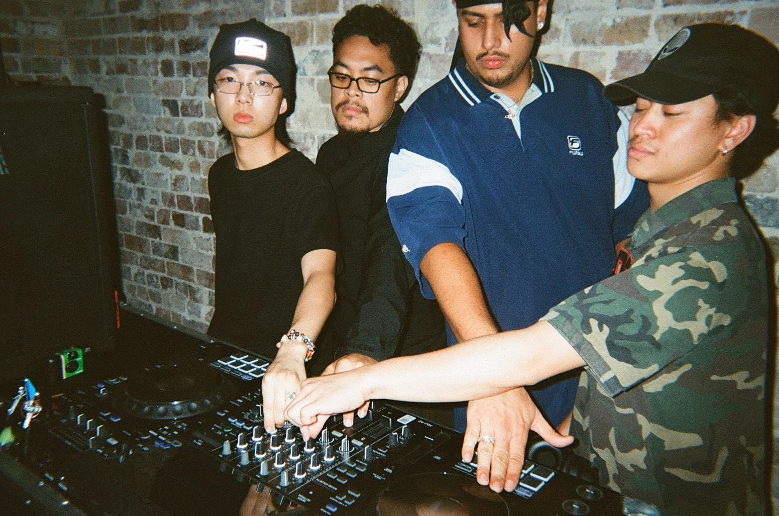 Four young men standing behind DJ equipment in a room with brick walls, with two of them manipulating the controls on a DJ mixer.