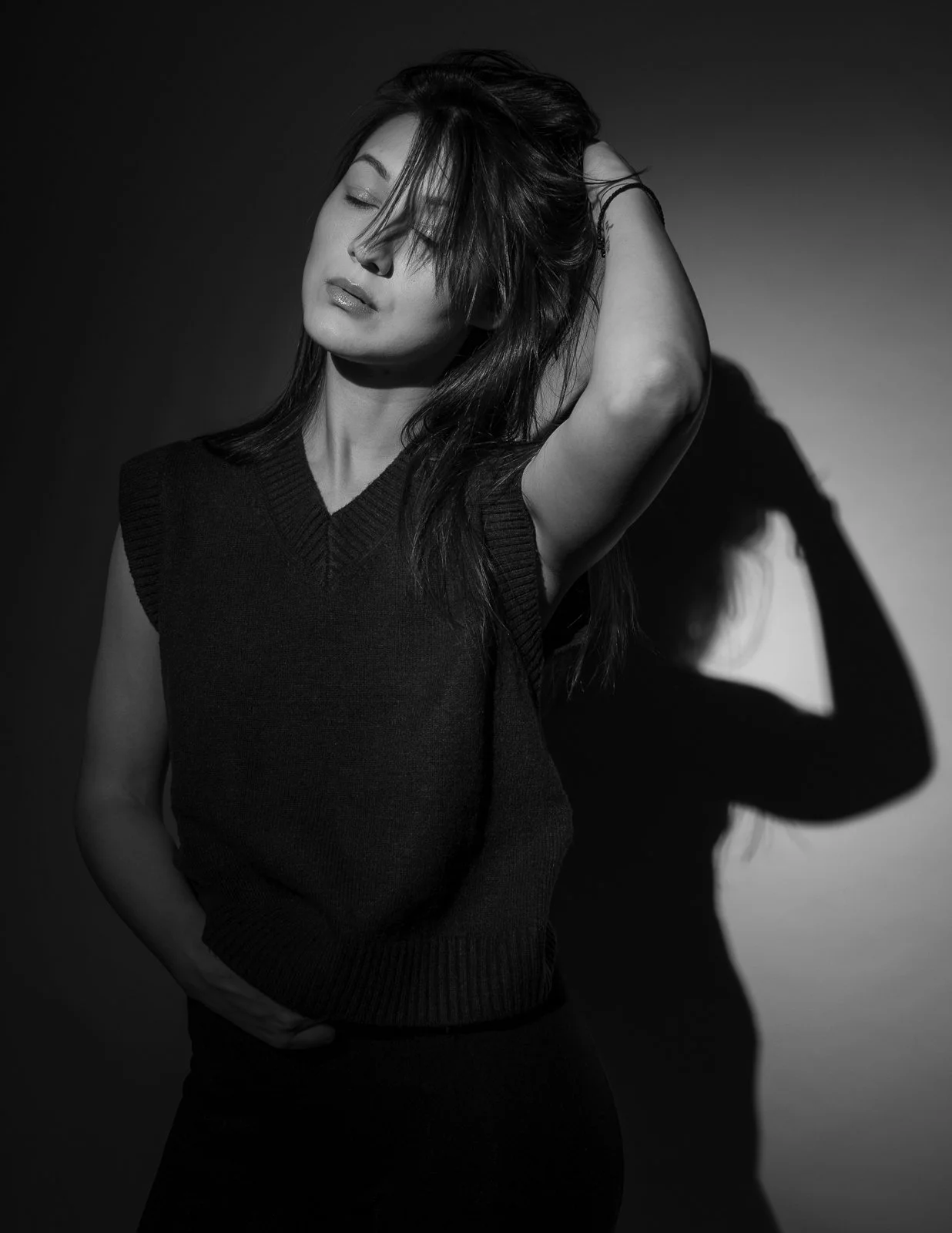 A woman with long hair and closed eyes, wearing a sleeveless sweater, posing with one arm raised, her hand on her head, in black and white with a shadowed background.