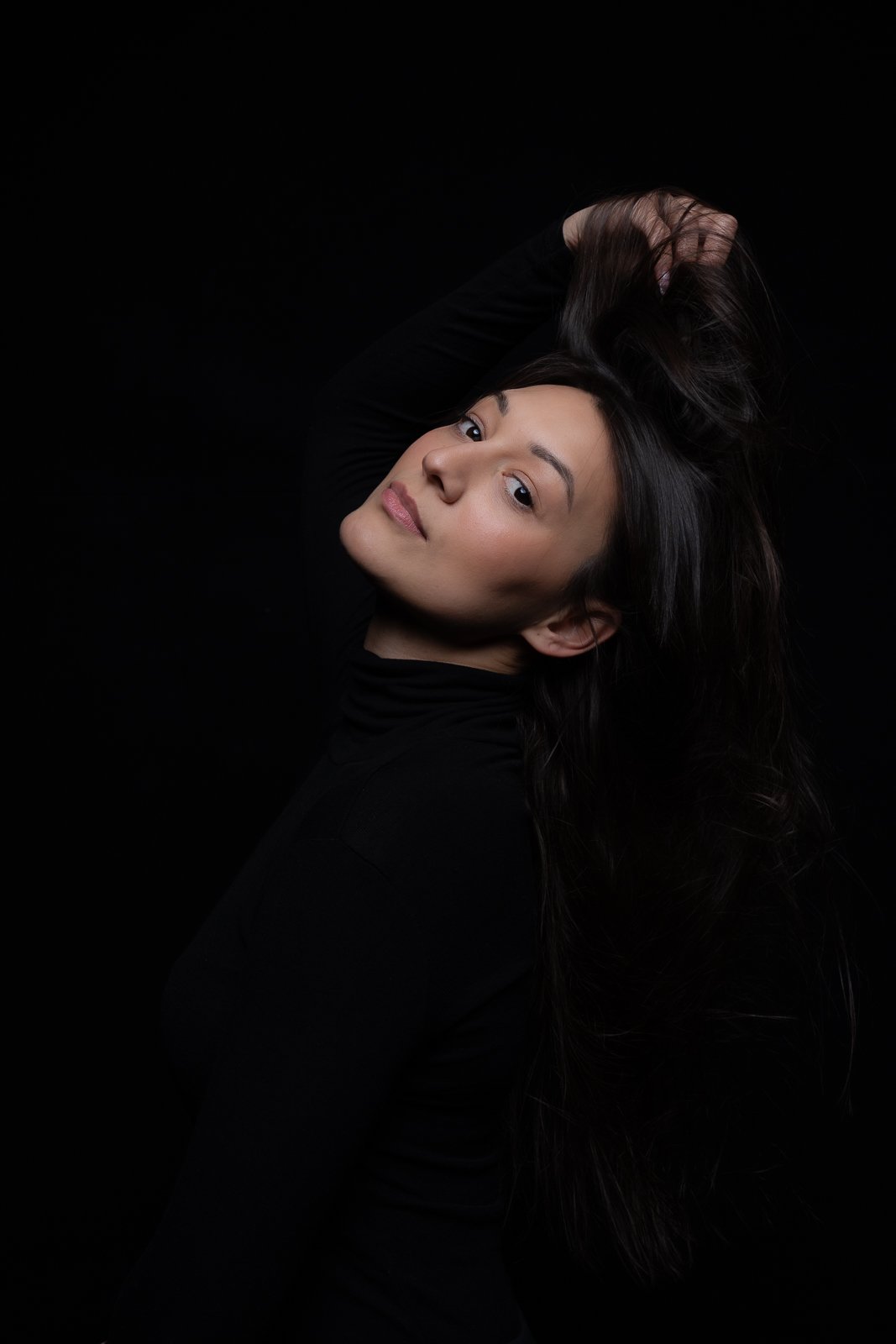 Portrait of a woman with long dark hair, wearing a black top, posing against a black background.