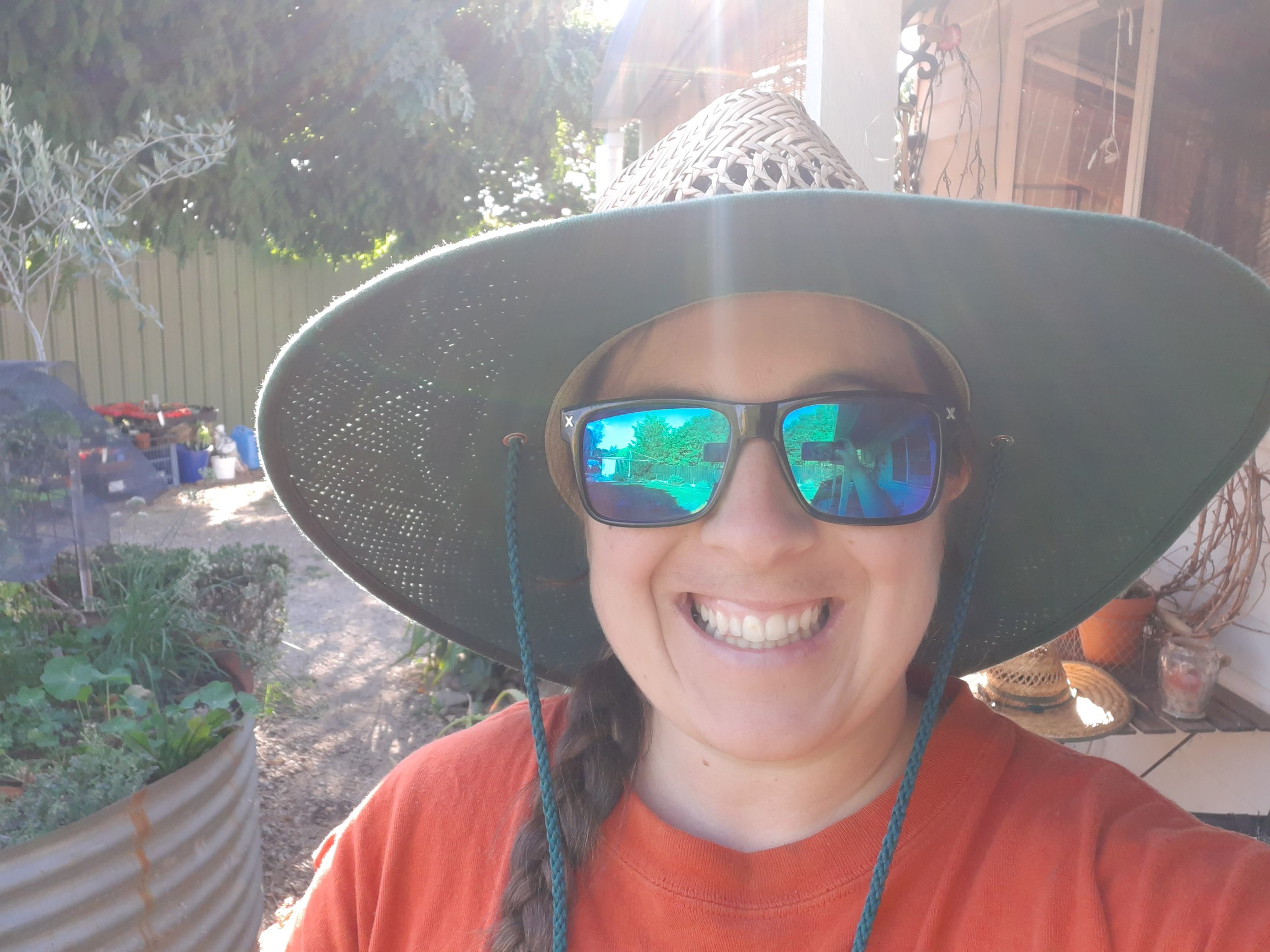 A woman wearing sunglasses and a wide-brimmed hat is smiling outdoors. The reflection in her sunglasses shows trees and a blue sky. She is wearing an orange shirt, and the background includes potted plants, garden tools, and outdoor furniture.