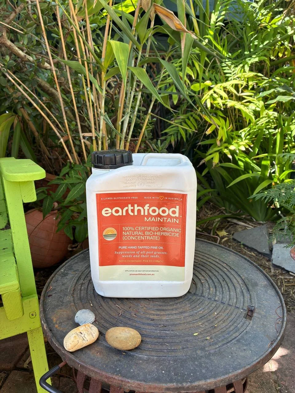 A white container of Earthfood Maintain organic bio-herbicide on a round outdoor table in a garden surrounded by green plants and rocks.