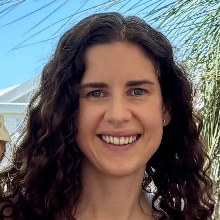Close-up of a smiling woman with curly brown hair outdoors, with blue sky and palm trees in the background.