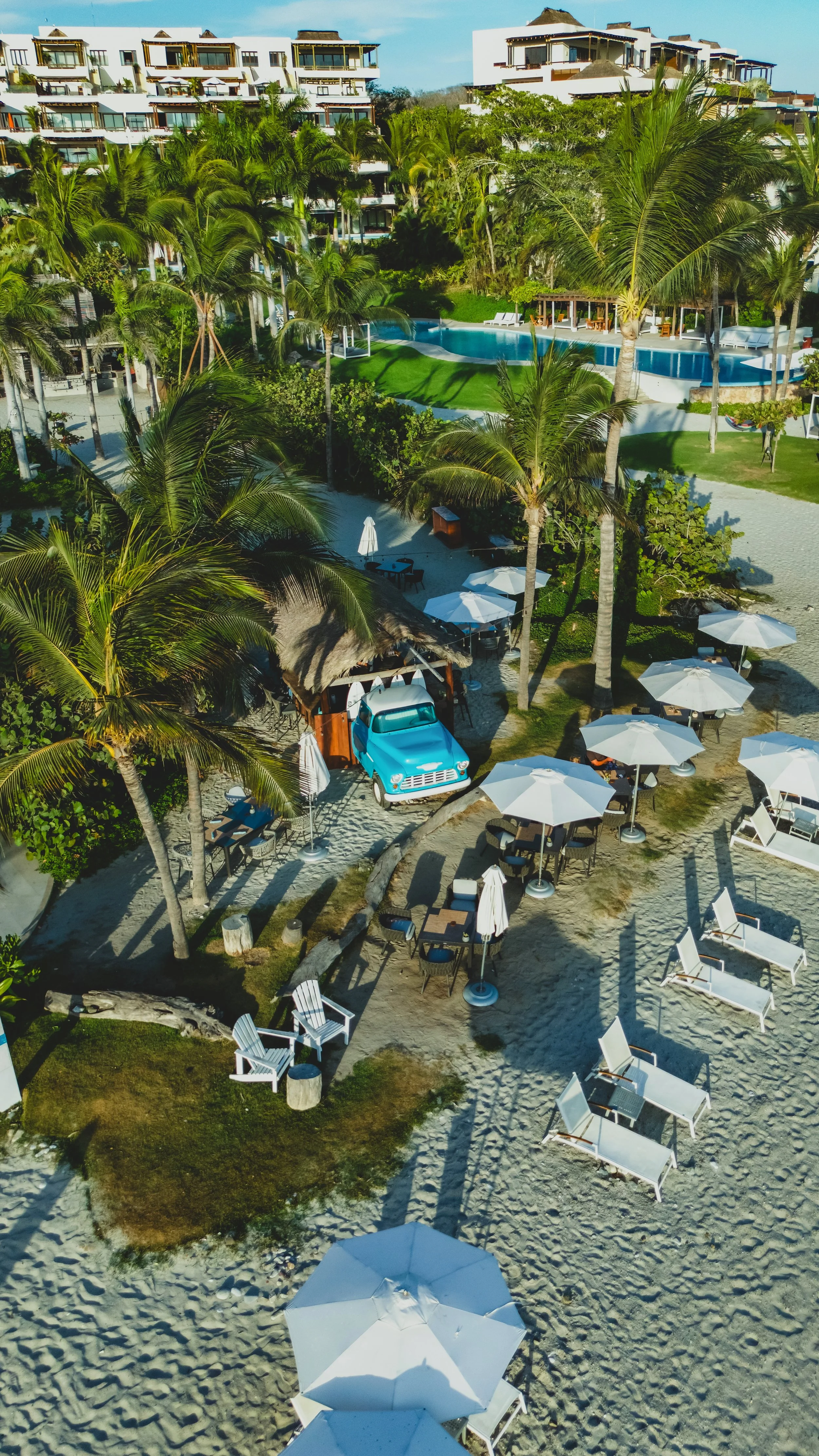 Aerial view of a tropical beach resort with white sand, lounge chairs, umbrellas, palm trees, a small hut, vintage blue truck, lush greenery, and a large swimming pool surrounded by modern buildings.