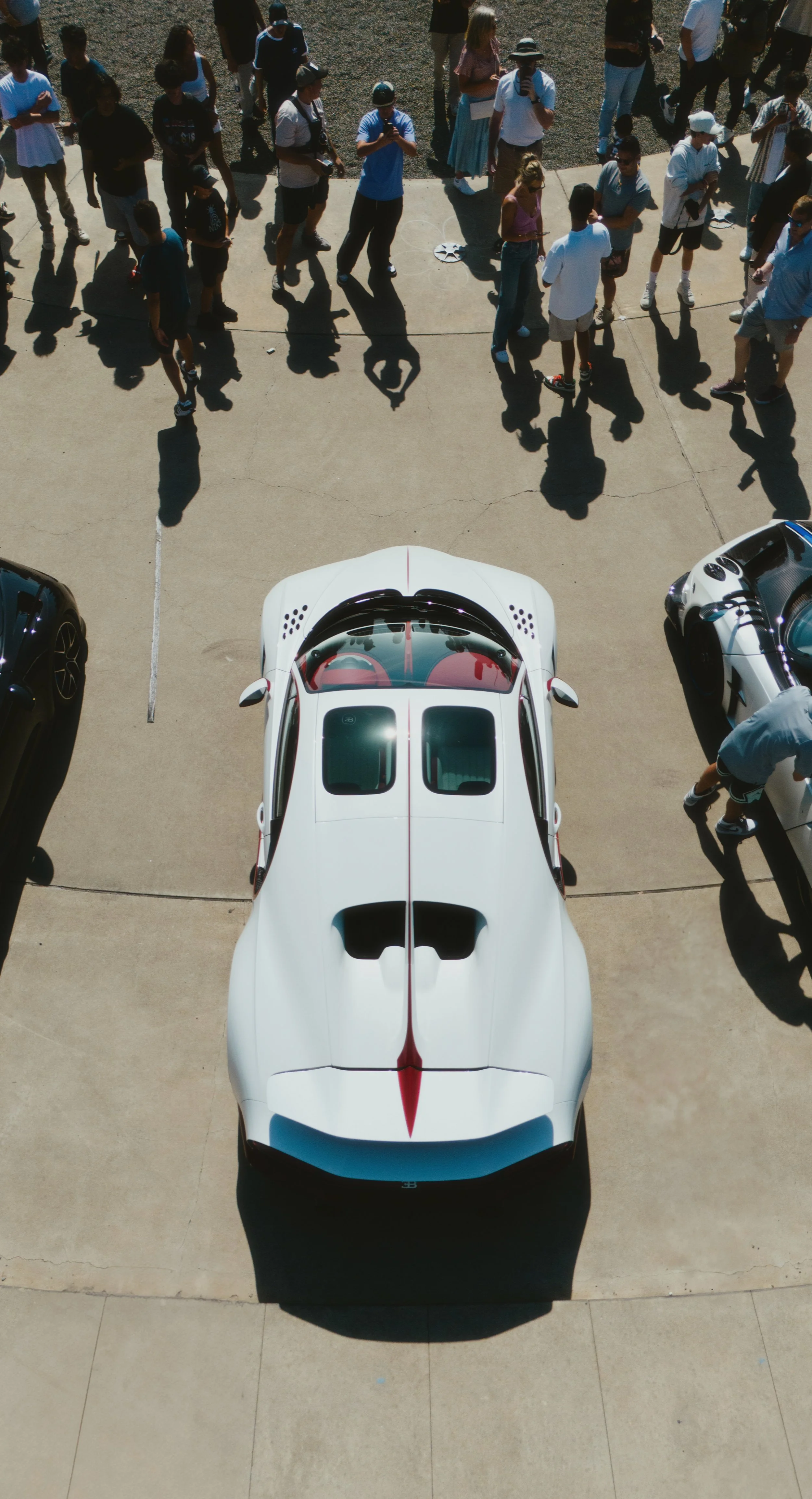 A white sports car with red accents viewed from above, parked on a concrete surface, with a crowd of people gathered around and looking at it.