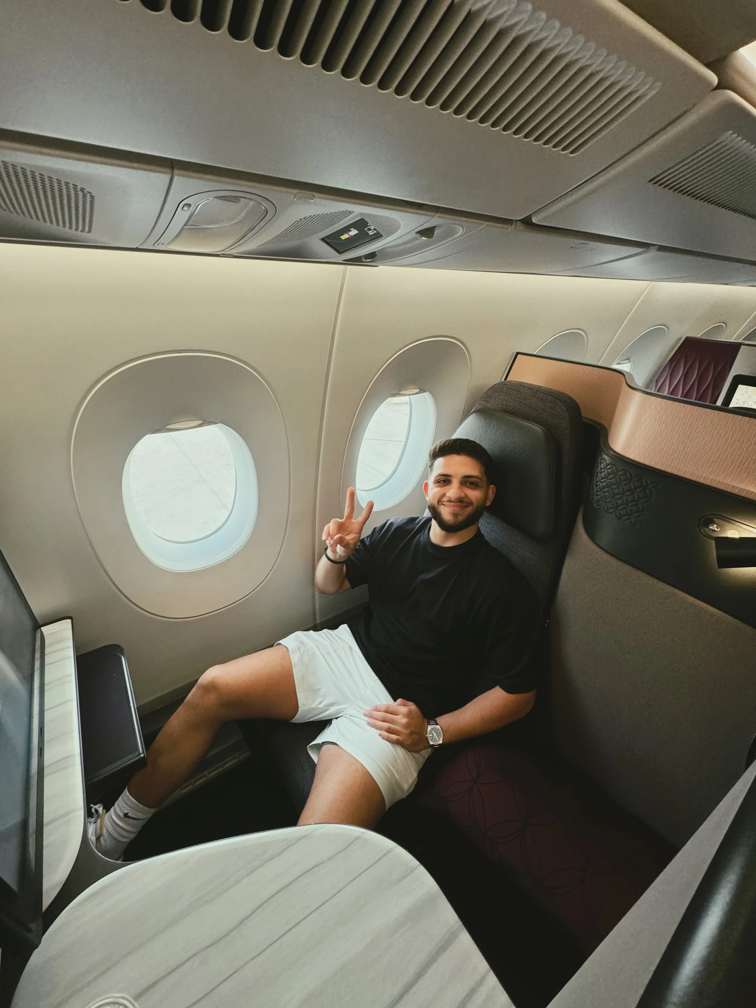 A man sitting in a business class airplane seat, making a peace sign and smiling, with windows and overhead compartments visible.