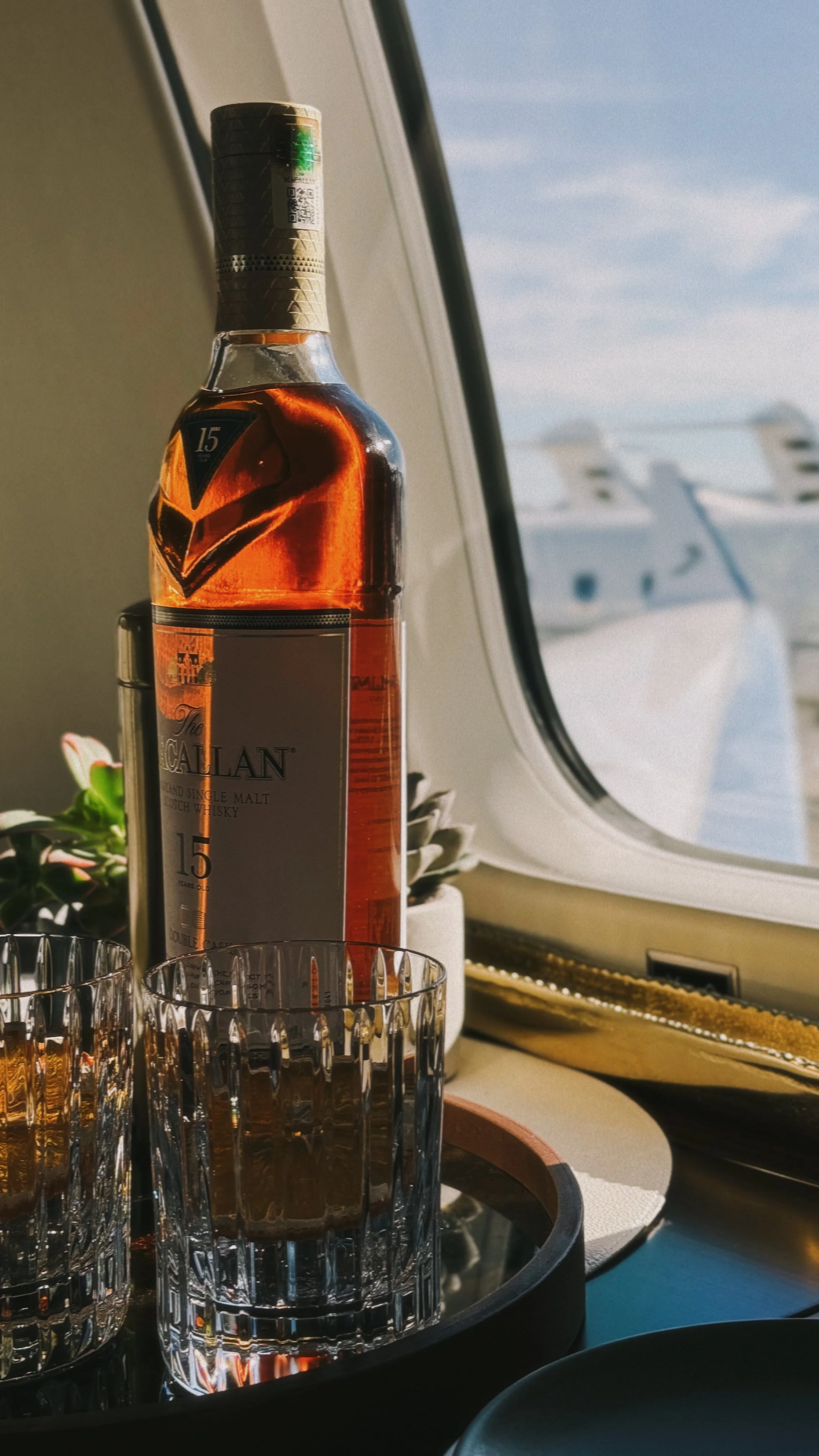 A bottle of The Glenlivet 15-year-old single malt Scotch whisky on an airplane window ledge with two empty glasses and a small plant, with yachts visible outside the window in the background.