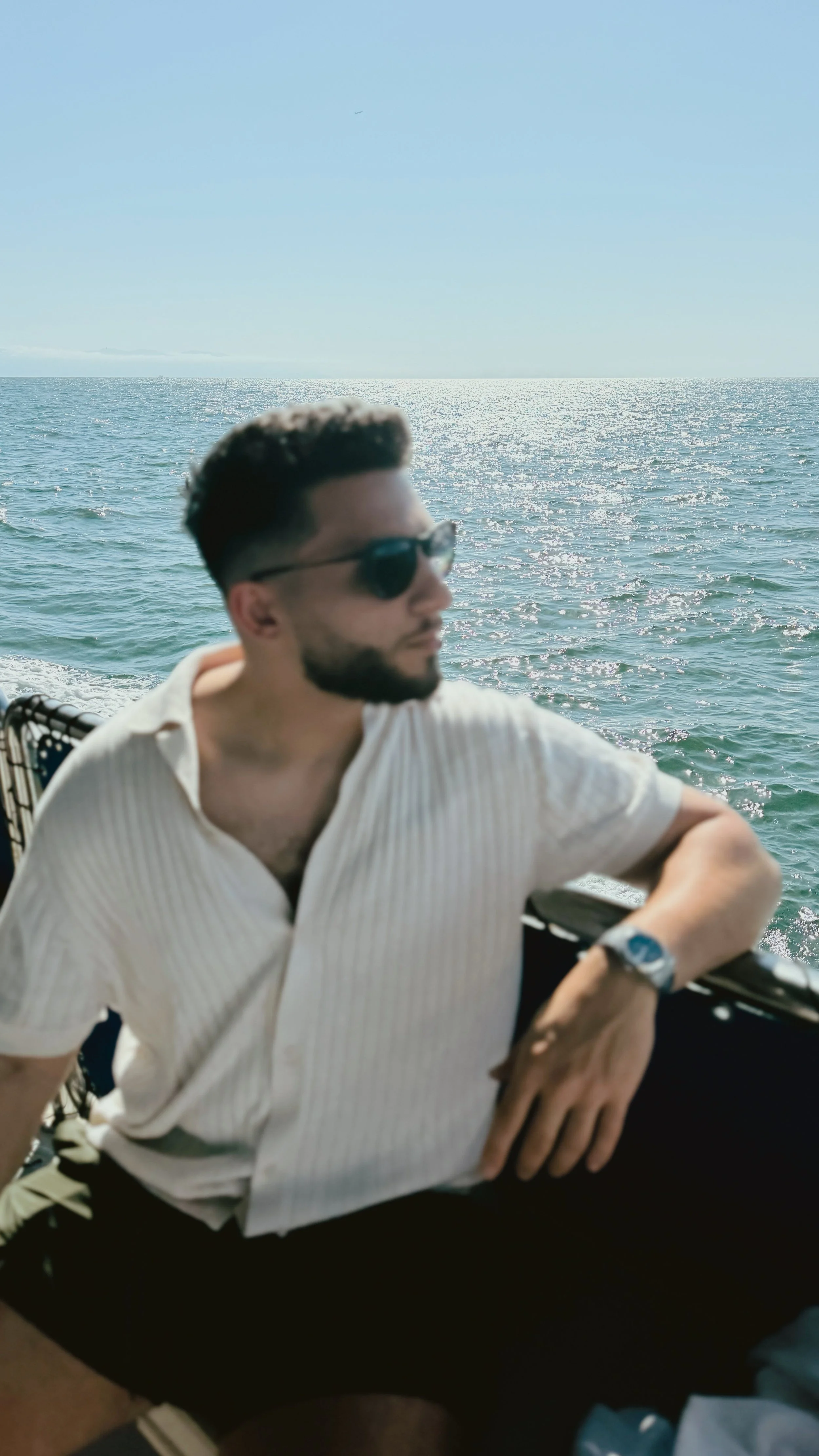 Man sitting outdoors near water, wearing sunglasses and a white shirt, with a background of the ocean and sky.