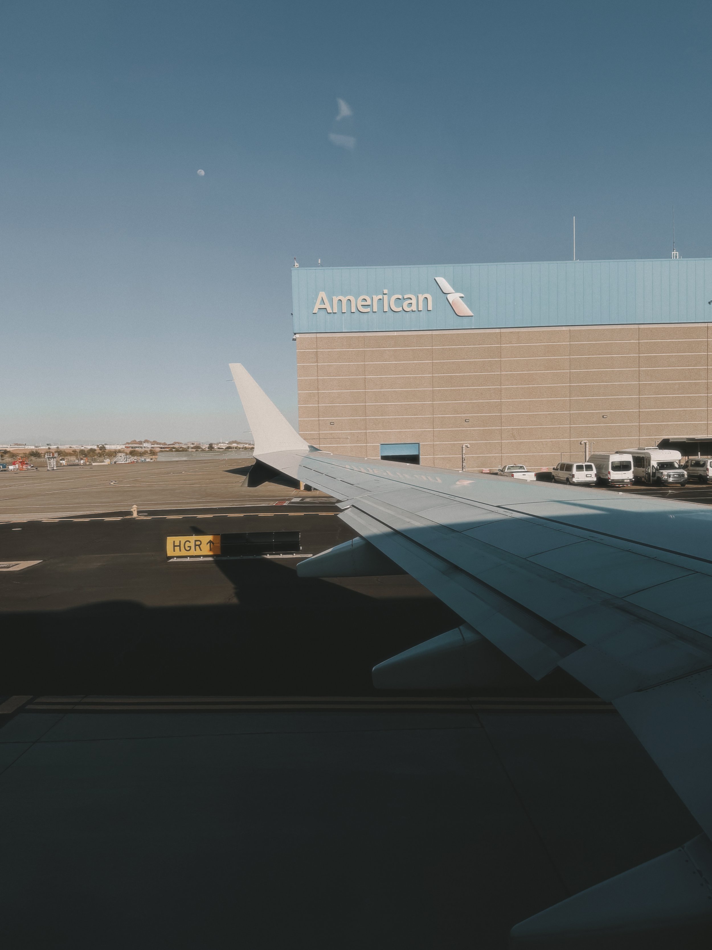 View of an airplane wing at an airport with the American Airlines terminal in the background showing the logo and some parked vehicles.