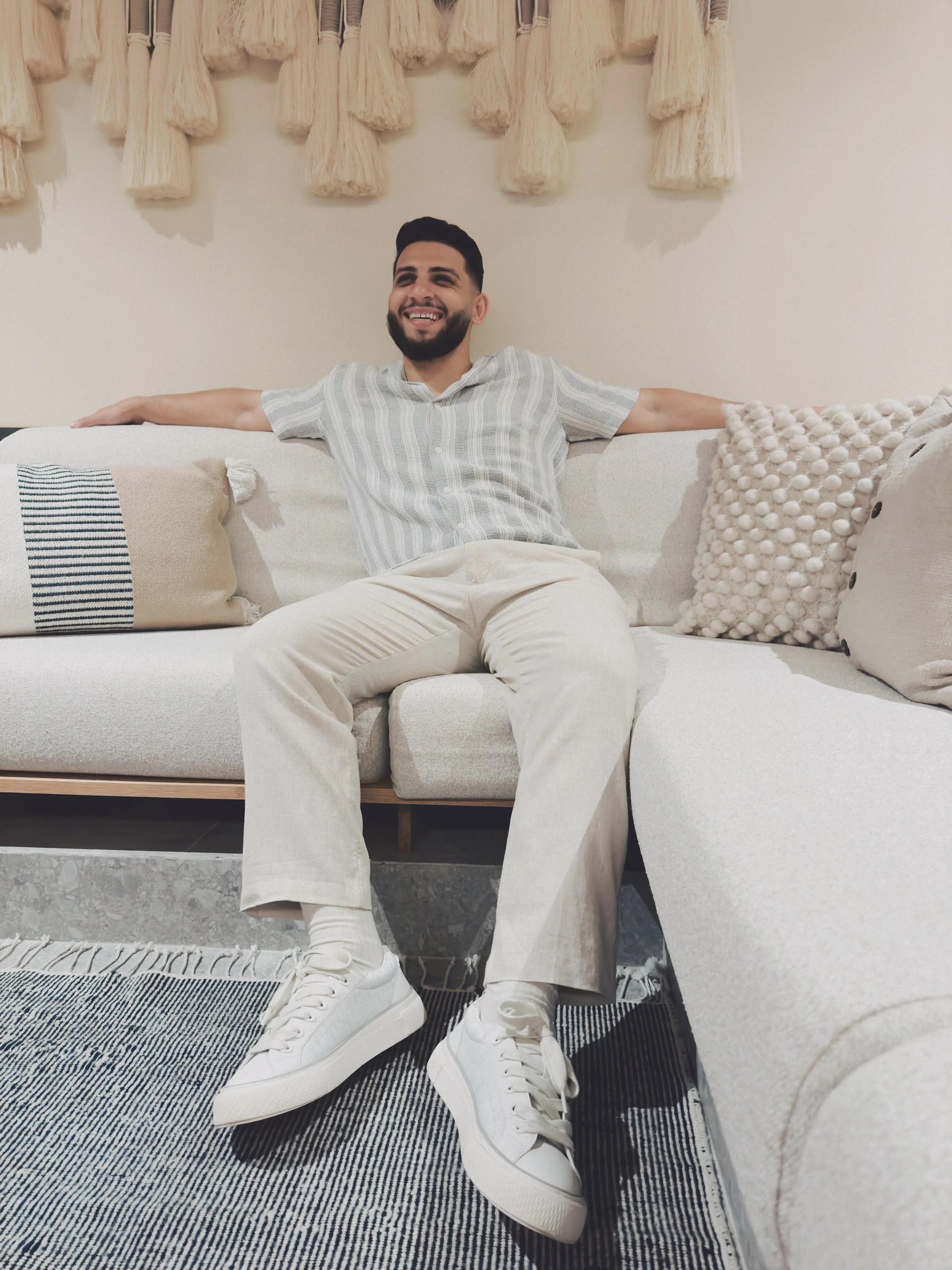 Ali Chokr on a light-colored sofa with his arms stretched out, smiling, in a cozy, well-decorated living room.