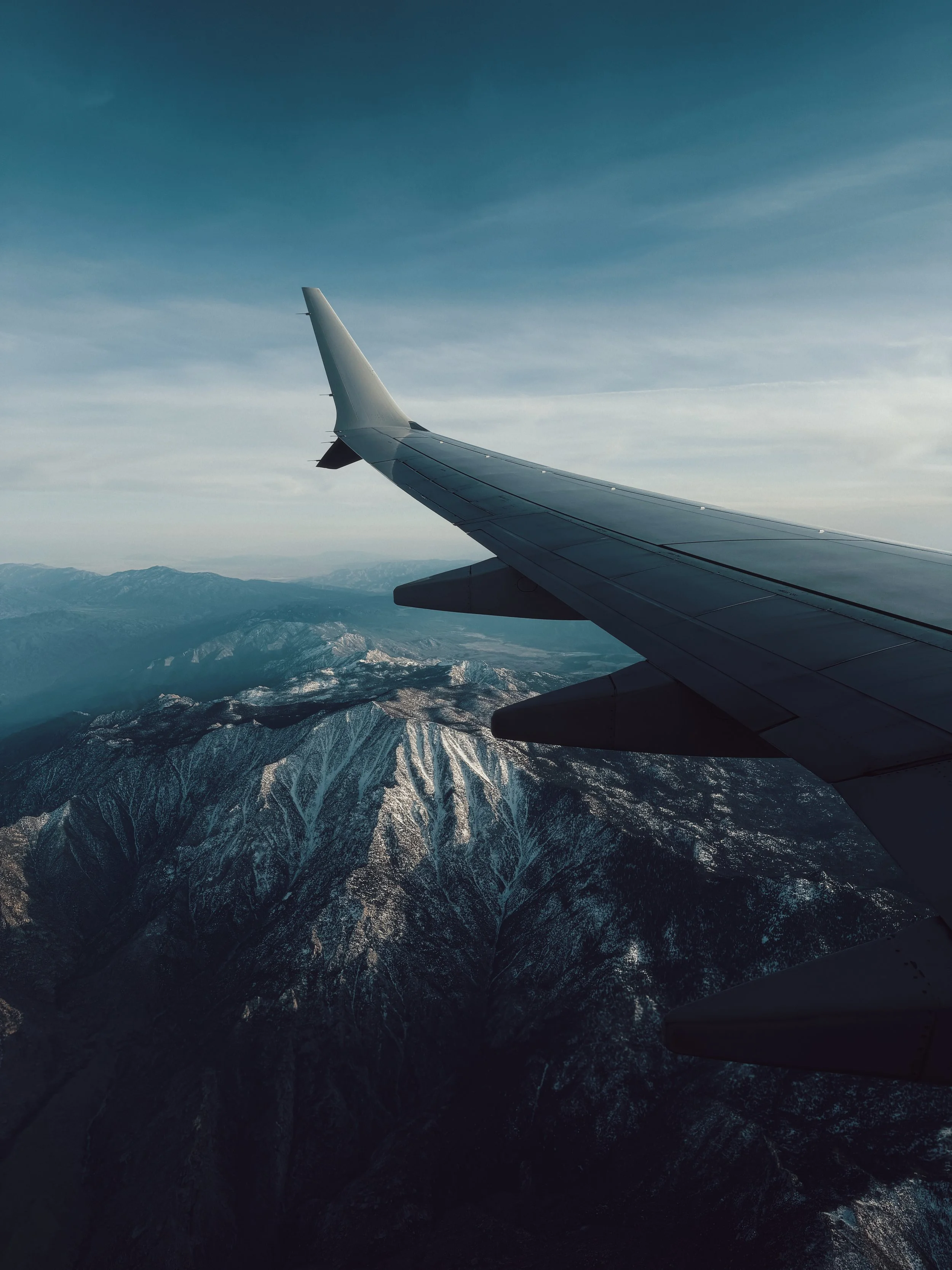 View from an airplane window showing an airplane wing and snow-capped mountain range below during flight.