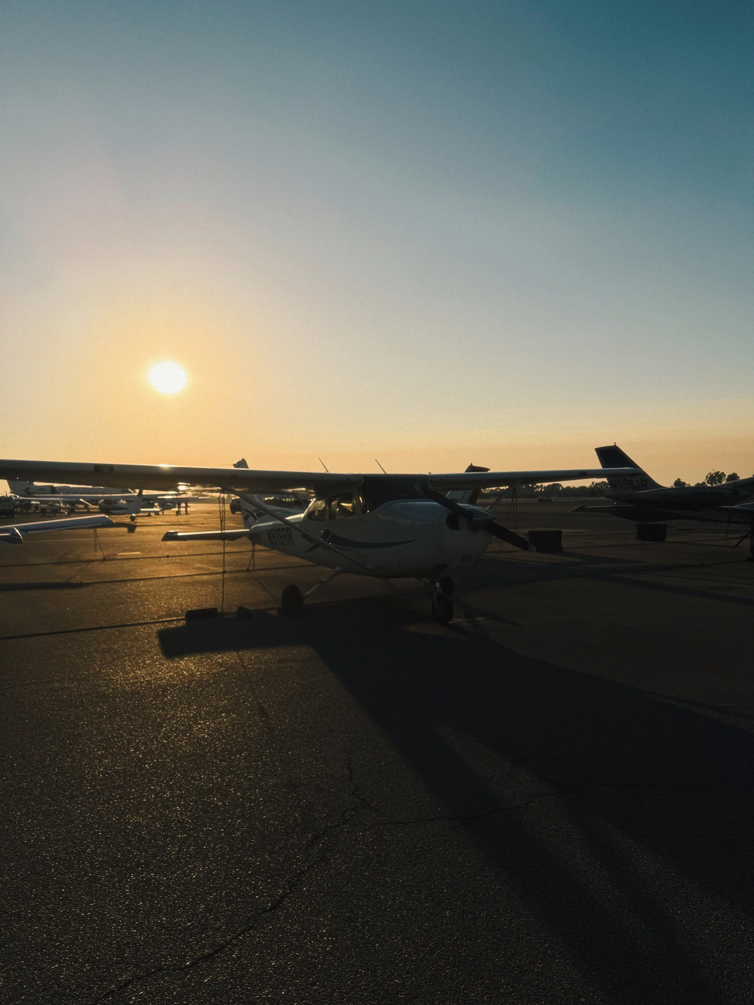Small airplane parked on tarmac at sunset with long shadows, other aircraft in background.