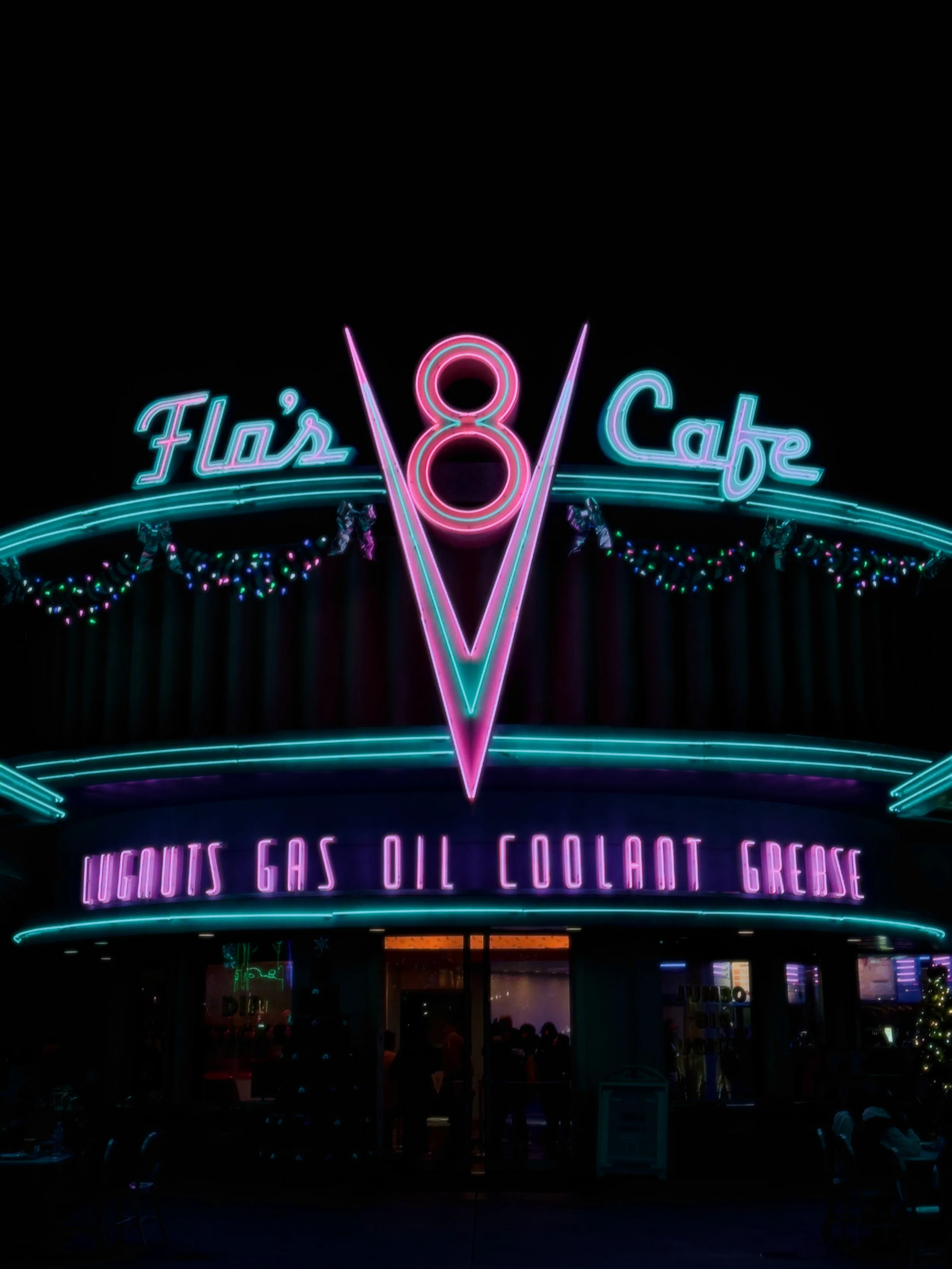 Neon sign for Flos Cafe with the words "Liquids Gas Oil Coolant Grease" under the main sign, decorated with colorful string lights.