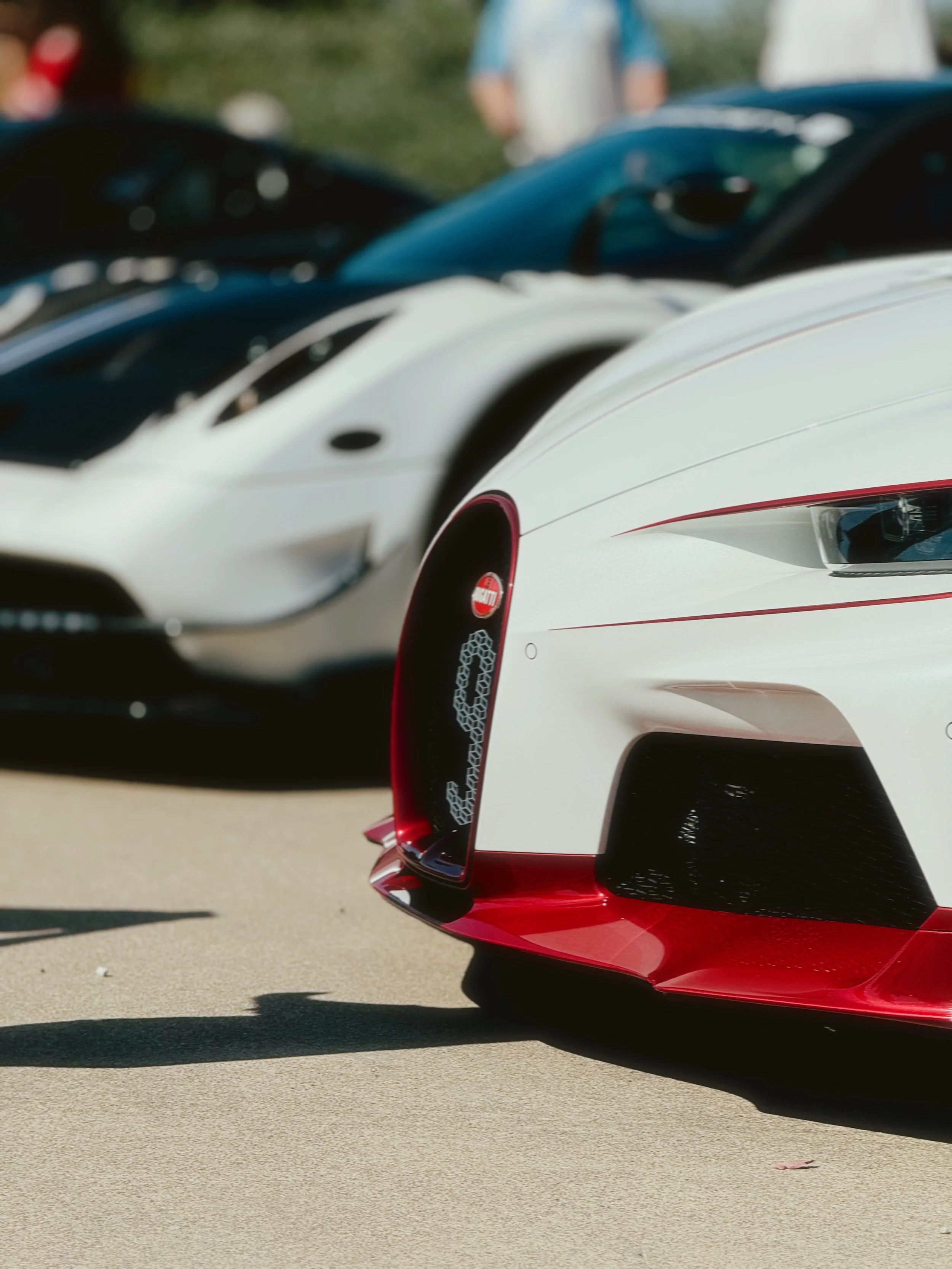 Close-up of the front end of a white and red sports car parked on pavement, with other cars and people in the background.